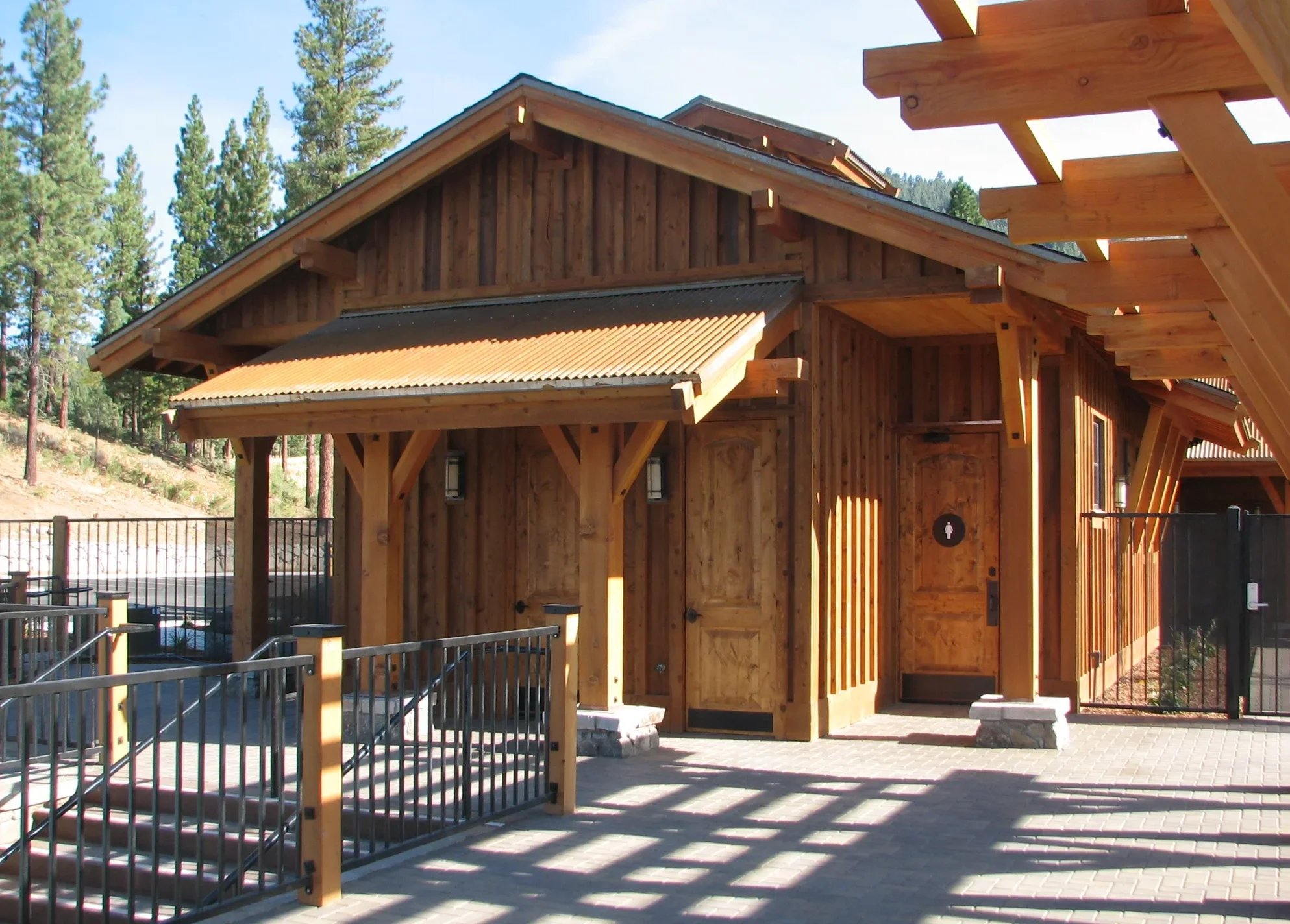 Wooden cabin with a sloped roof, located in a wooded area with tall pine trees, possibly a recreation area or park.