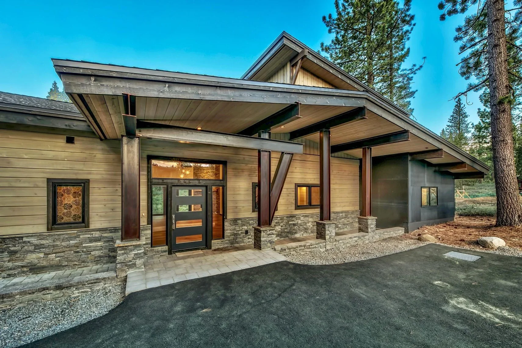 Modern house with wooden and stone exterior, black door, and large overhanging roof, surrounded by tall pine trees.