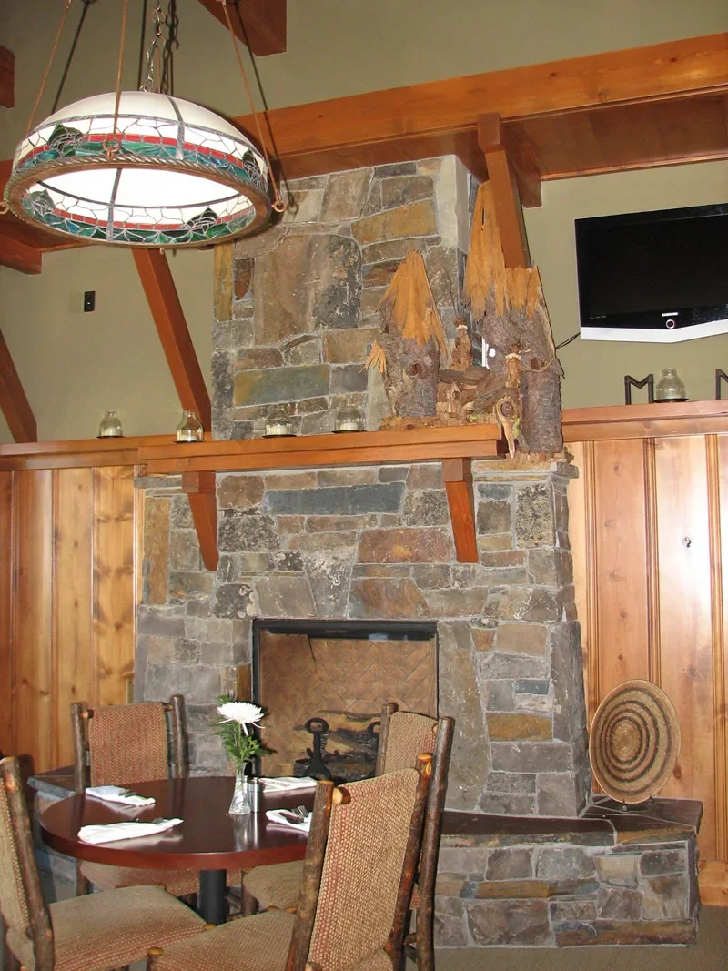 A cozy dining area with a round wooden table, four chairs, and a stone fireplace with a wooden beam mantel. There is a stained glass pendant light hanging from the ceiling, and rustic wood decor on the mantel. The space has wood paneling and a flat-s