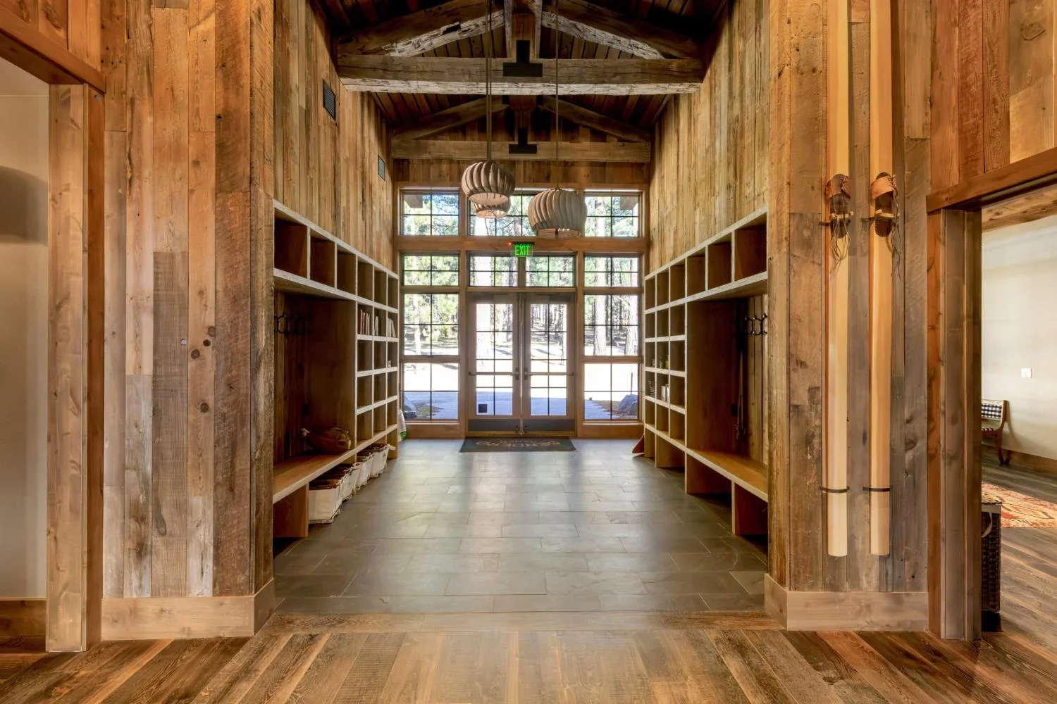 Vaulted lodge entry with reclaimed wood walls, exposed timber trusses, sculptural pendant lights, built-in benches and cubbies, slate tile flooring, and glass doors facing the pine forest.