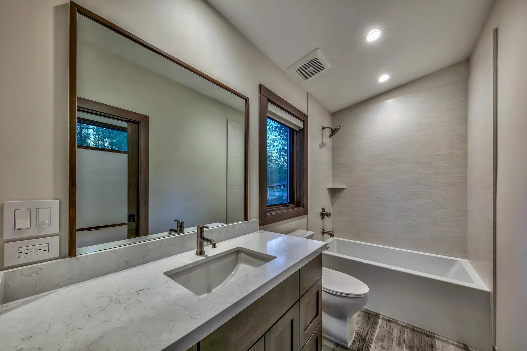 Modern bathroom with vanity, sink, large mirror, window, bathtub, and neutral-colored walls and flooring.