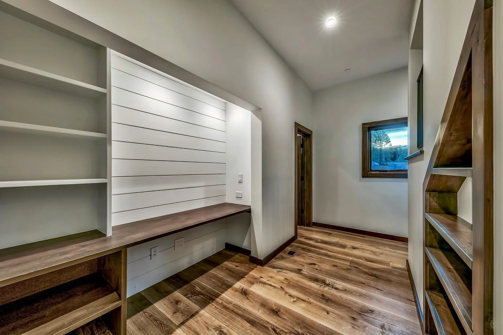 Empty room with white walls, wooden floors, and built-in shelves and a countertop along the walls. There is a small window with a view of trees outside.