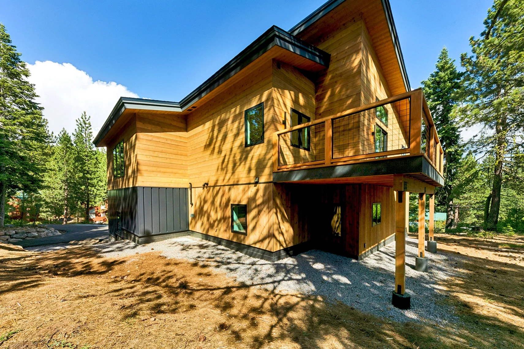 Modern two-story wood-clad home nestled among evergreen trees, featuring a sloped roof, large windows, and a natural forest setting.