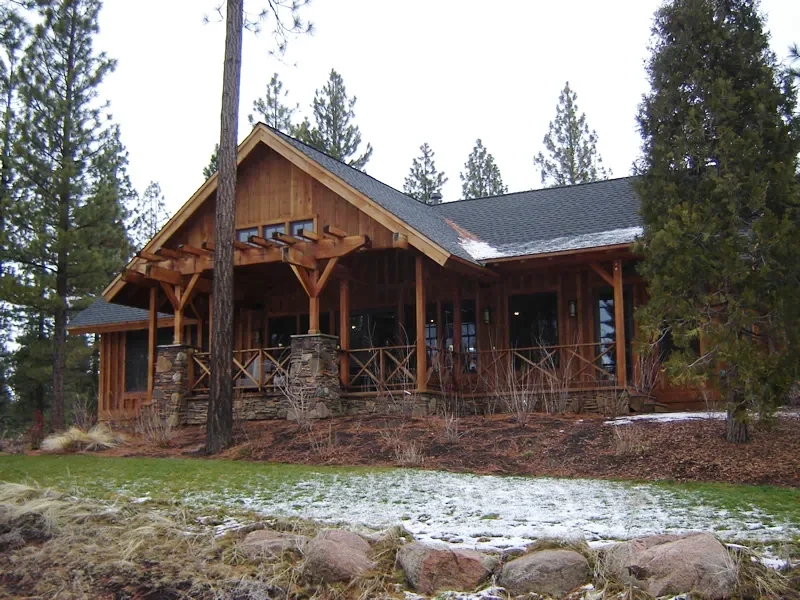 A large wooden house with a stone foundation, surrounded by trees in a forested area with patches of snow on the ground.
