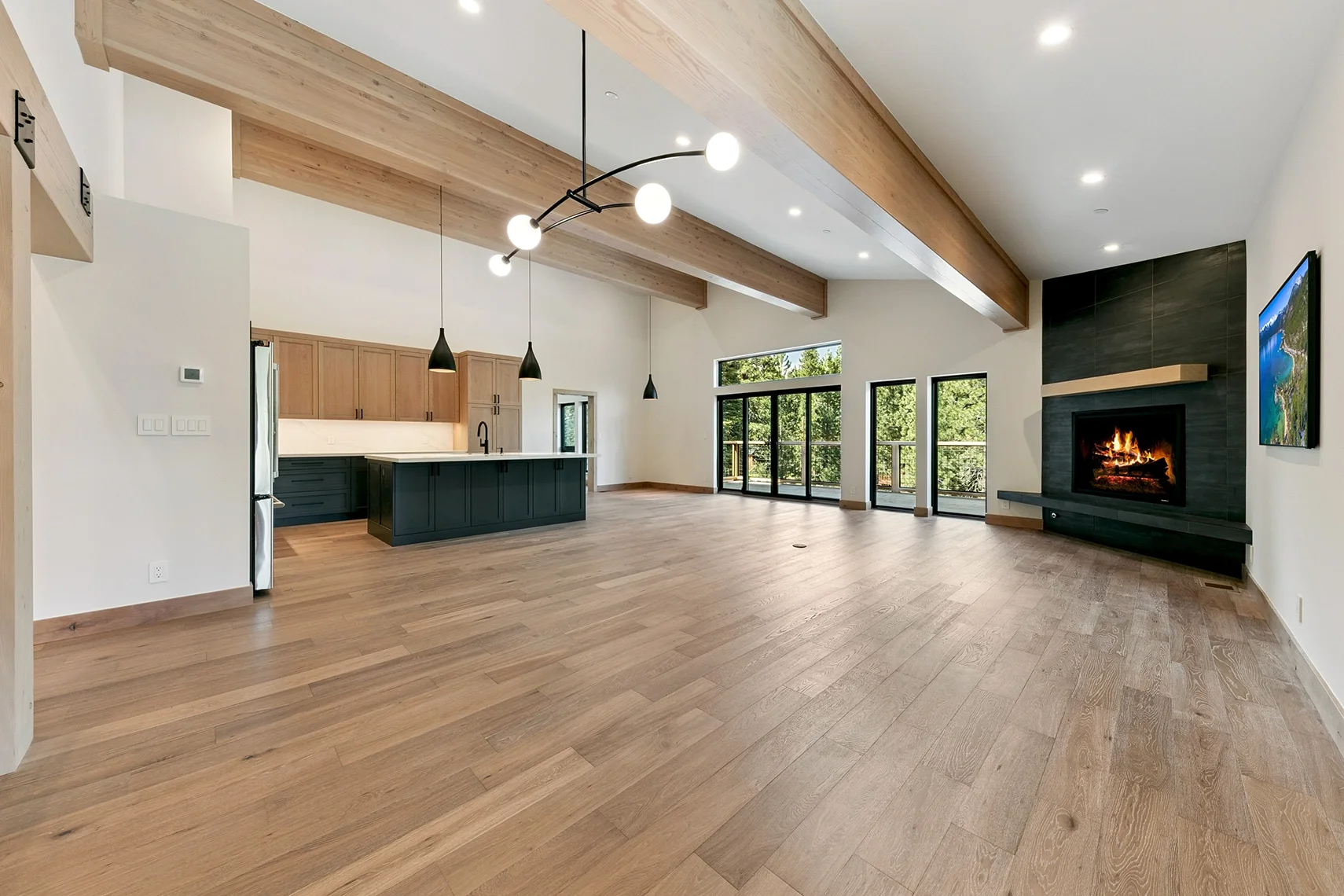 Open-concept modern mountain home interior featuring a kitchen with dark cabinetry, a spacious living area, natural wood flooring, and a dramatic black stone fireplace.