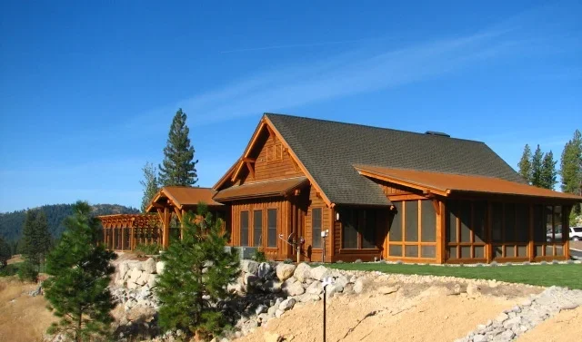 A large wooden house with a steep black roof and a screened porch, surrounded by trees and rocks under a clear blue sky.