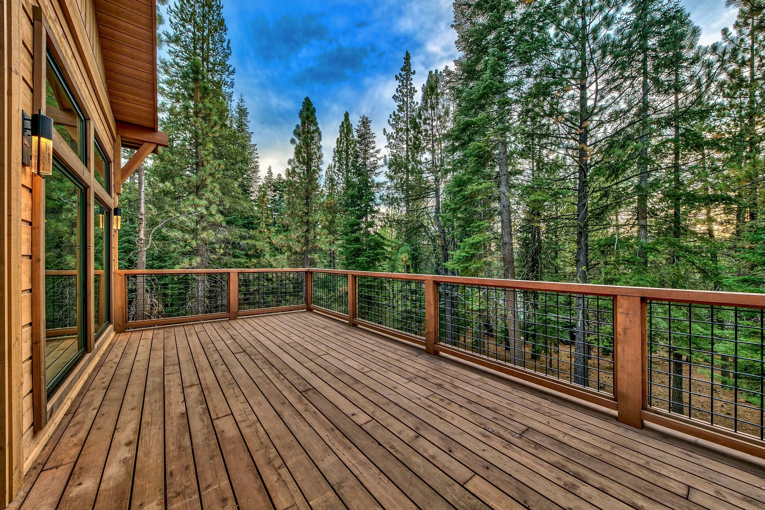 A spacious wooden deck attached to a house, overlooking a dense forest of tall green pine trees under a partly cloudy sky.