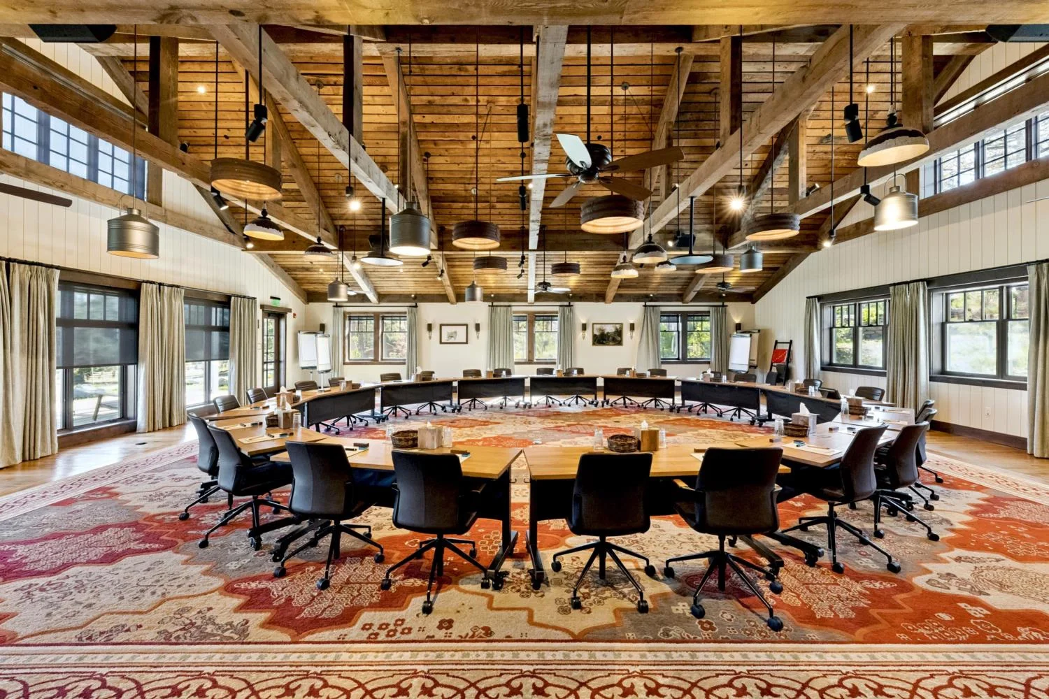 Large conference room with desks arranged in a circle, rolling chairs, an oversized patterned rug, exposed timber beams, varied pendant lights, and rows of windows.