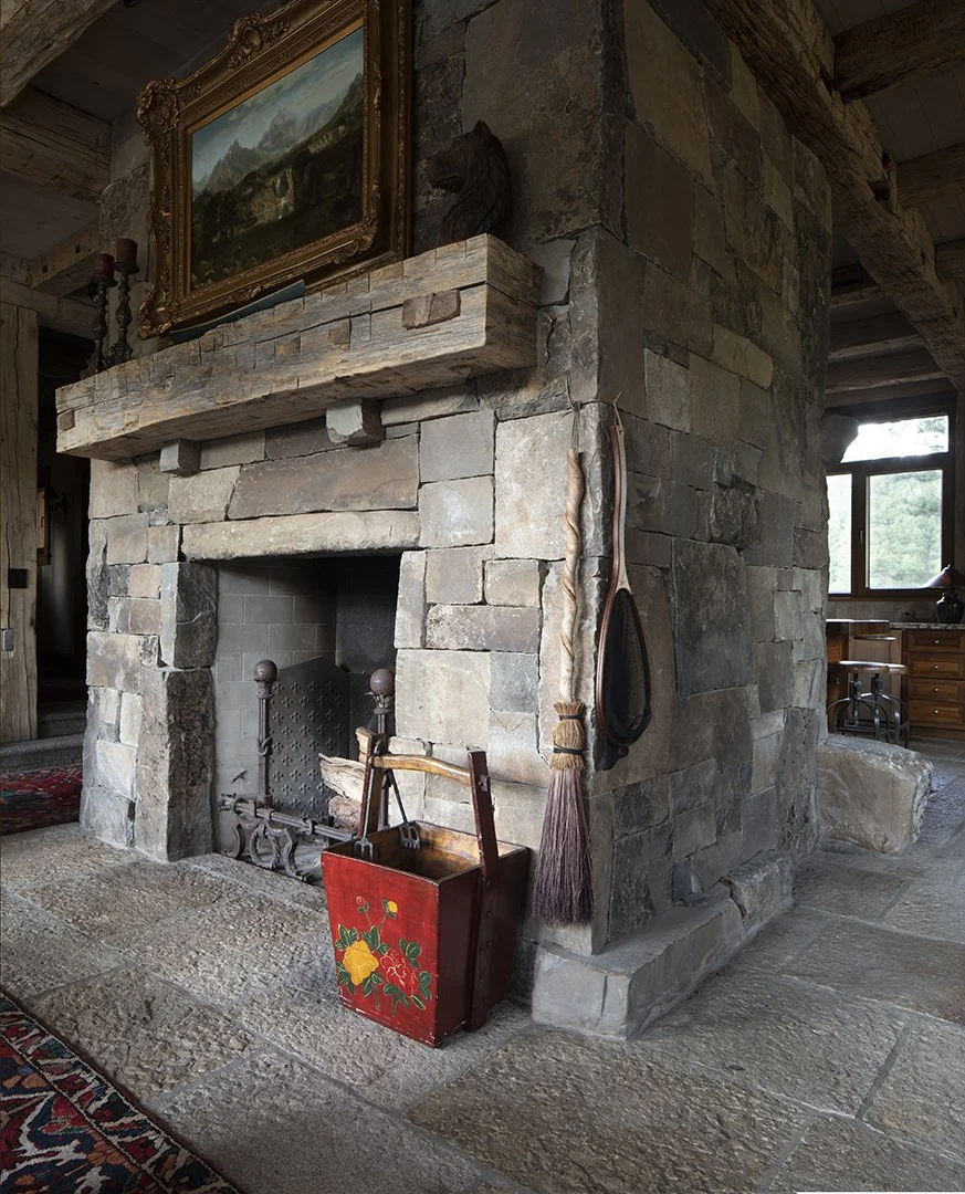 A rustic stone fireplace with a wooden mantel, next to a broom and a red decorative bucket with floral patterns. There is a painting above the mantel and a bear head mounted on the wall. The room has wooden beams and a window in the background.