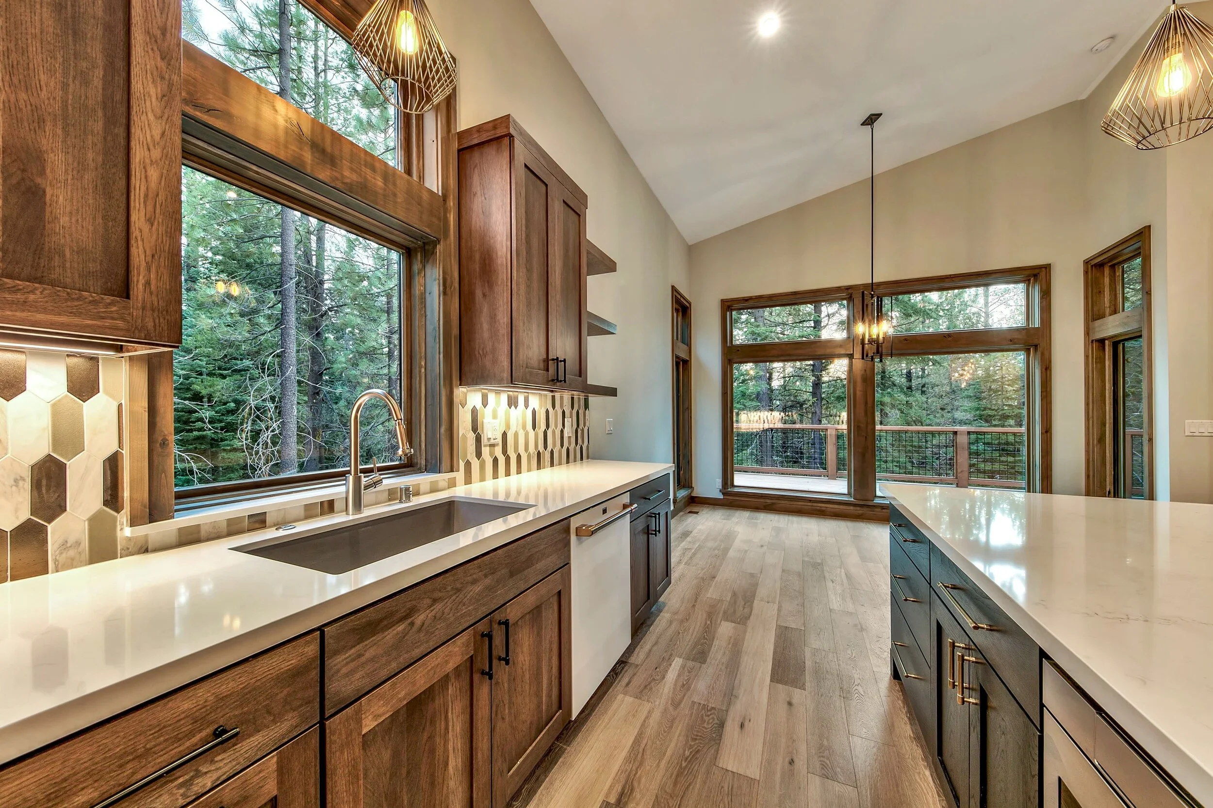 Modern kitchen with wooden cabinets, a white countertop with a black sink, ceramic tile backsplash, large windows showing trees outside, and hardwood flooring.