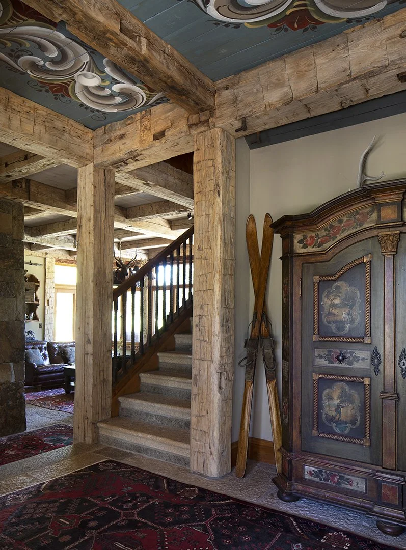 Interior of a rustic home with wooden beams, a staircase, vintage furniture, and vintage skis leaning against the wall.