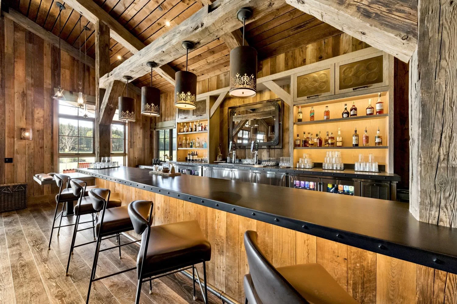 Modern mountain lodge bar with reclaimed wood paneling, a black metal counter, leather stools, illuminated shelving, decorative pendant lights, and exposed timber beams.