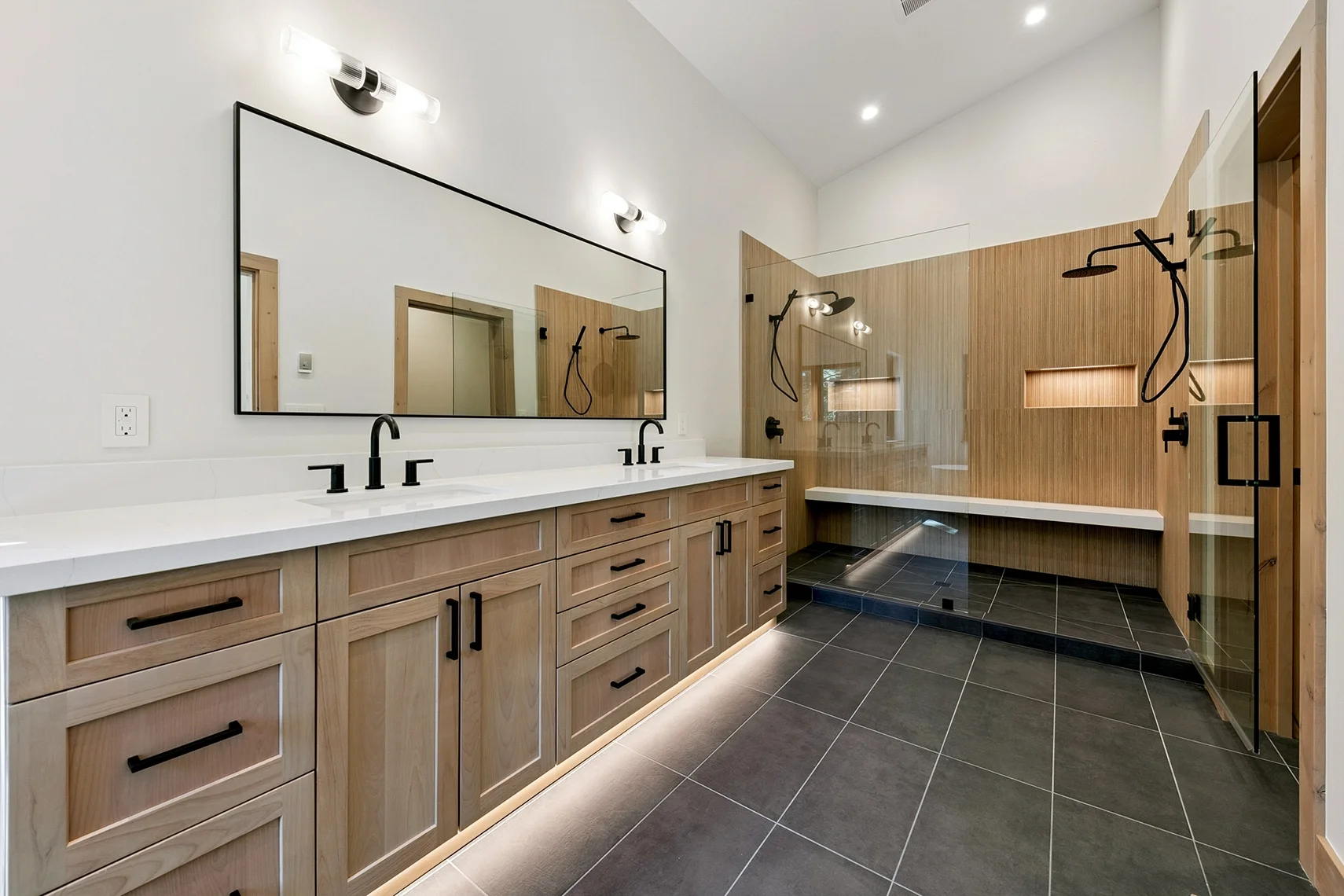 Modern bathroom with white walls, black tile flooring, a double vanity with light wood cabinetry, white countertops, a large mirror, and a glass-enclosed double shower with a built-in bench.