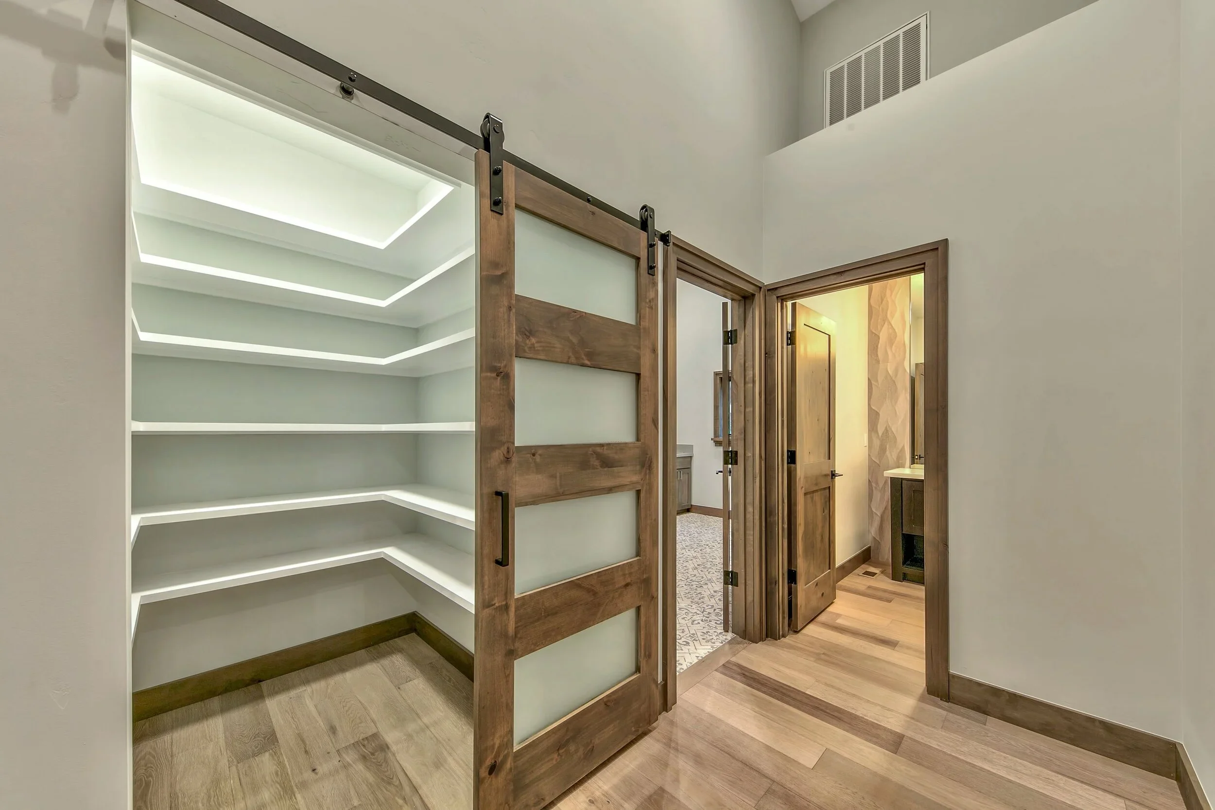 Interior of a house with a sliding barn door on a library or pantry with wooden shelves and a doorway leading to a bathroom with a vanity and mirror.