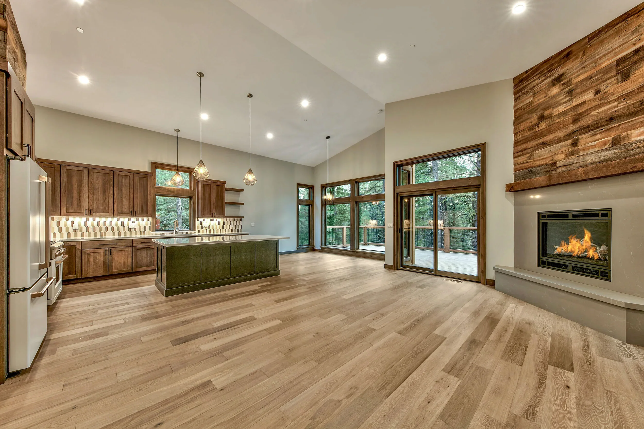 Open-concept living area with kitchen and fireplace, featuring wooden floors, large windows, and a deck outside.