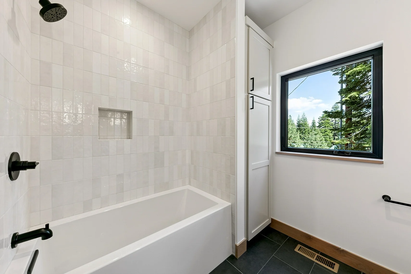 Modern white bathroom with a tiled bathtub and a window overlooking a scenic view of pine trees from above.