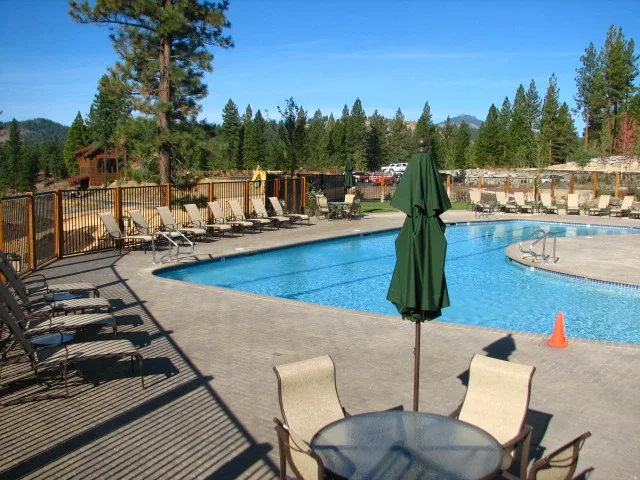 Empty swimming pool area with lounge chairs and an umbrella, surrounded by trees and mountains in the background.
