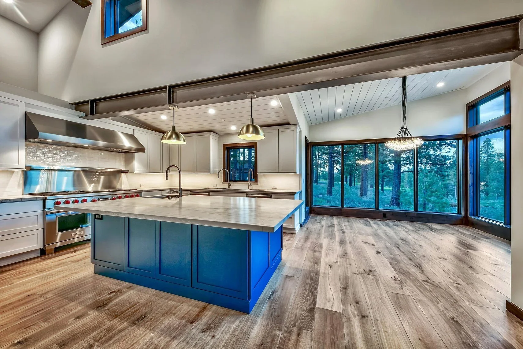 Open-concept kitchen with a large island, stainless steel appliances, white cabinetry, wood flooring, and large windows showing trees outside.