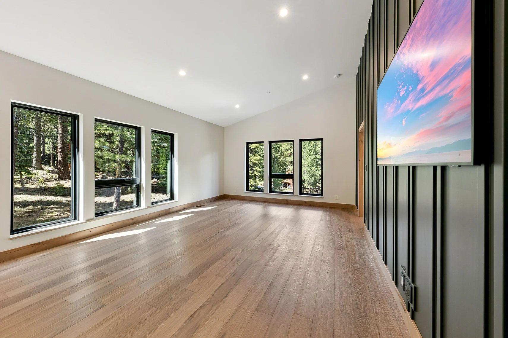 Spacious living room with white walls, natural wood flooring, five large windows, a black wood-paneled accent wall, and a large mounted TV.