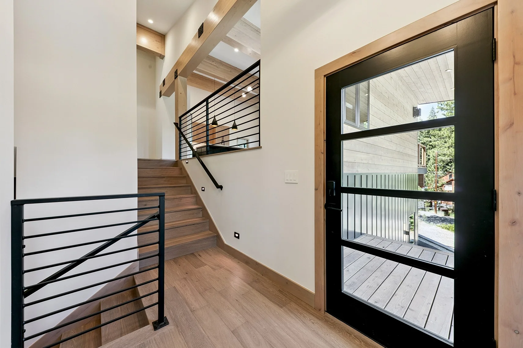 Modern mountain home entryway featuring natural wood flooring, exposed timber beams, horizontal iron stair railings, a floating staircase, and a black-framed glass front door.