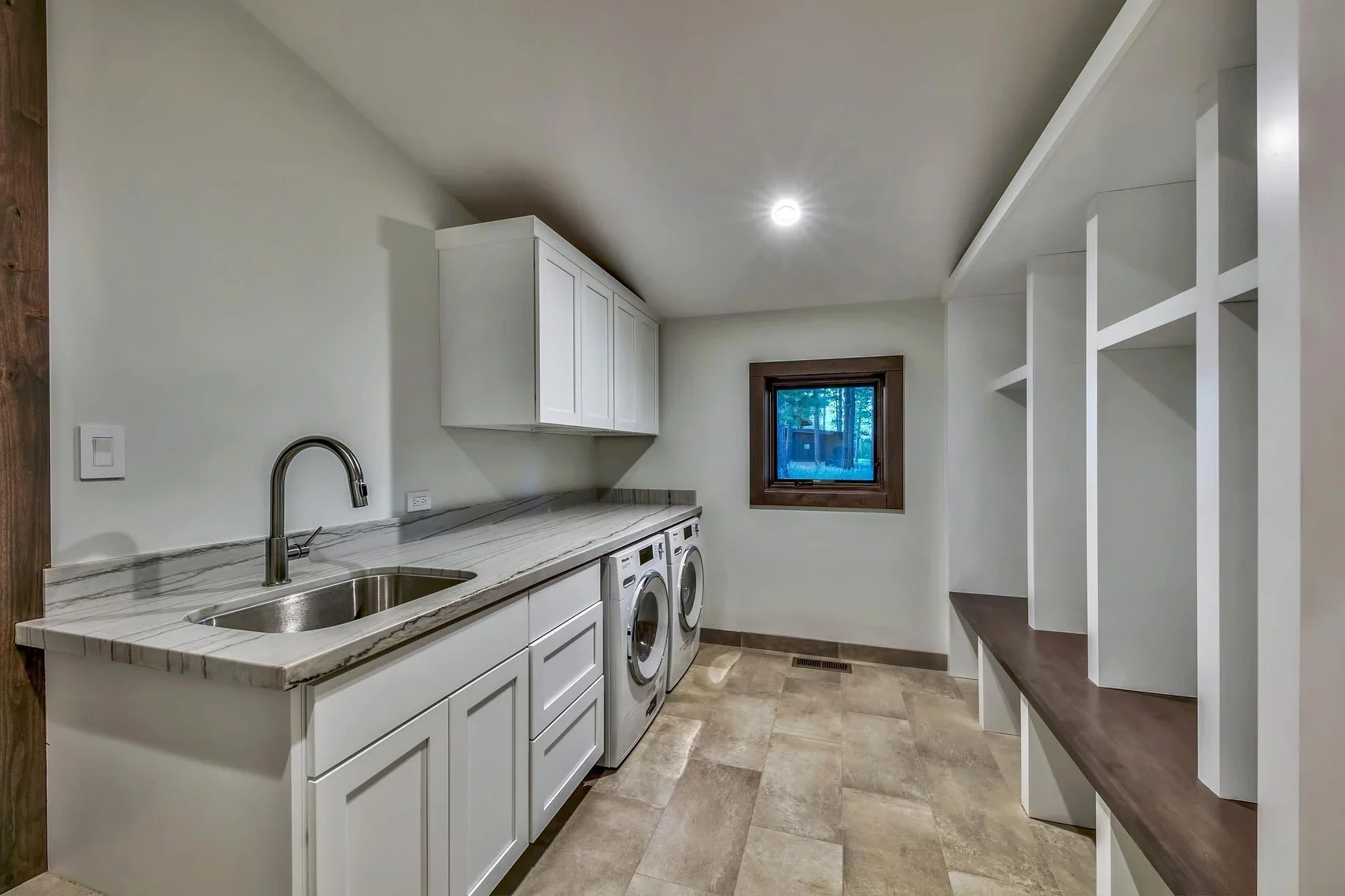 Laundry room with white cabinets, a marble countertop, double washer and dryer, a small window, and a wooden shelf unit.