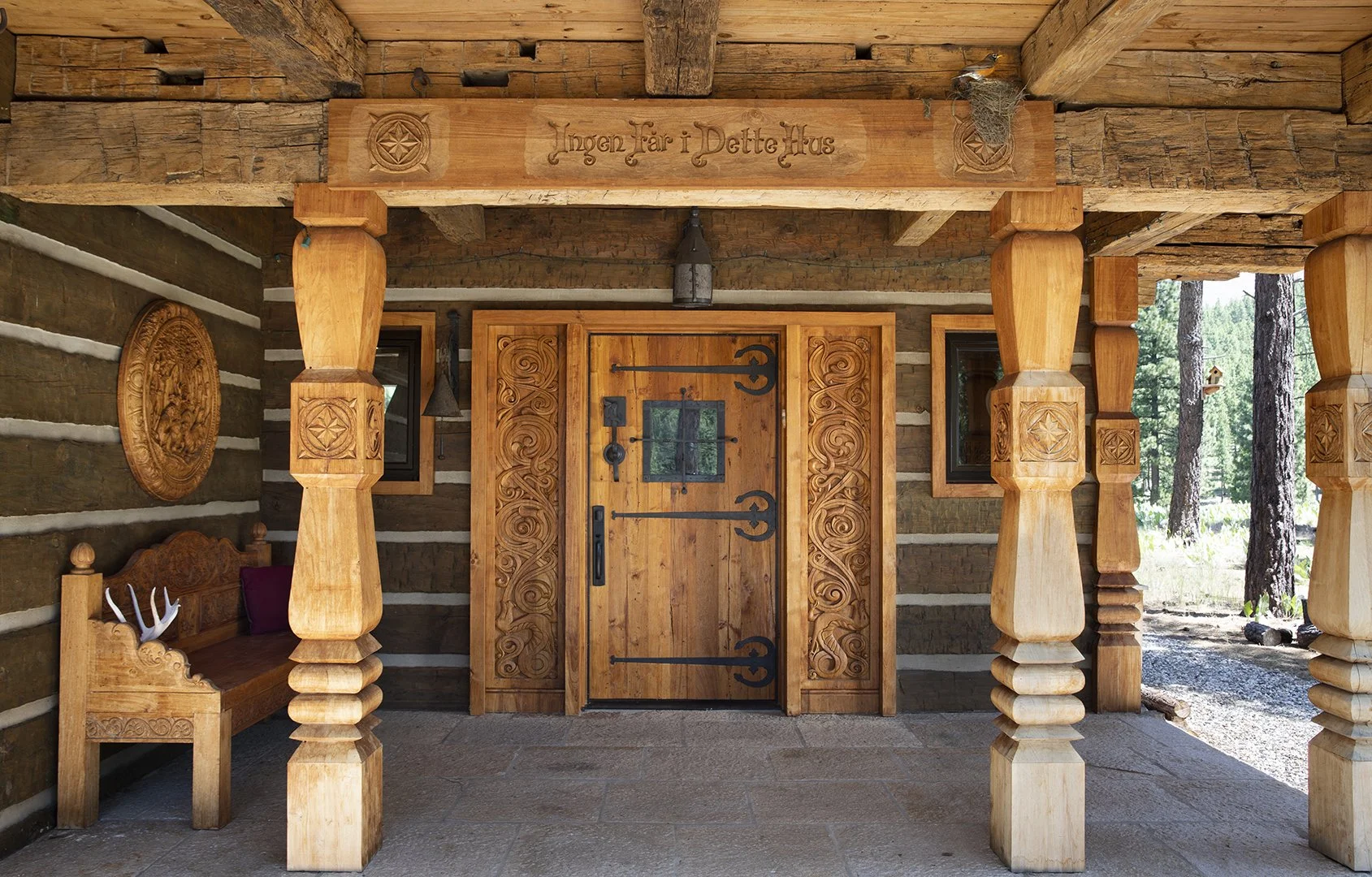 Wooden house entrance with carved door and decorative columns, surrounded by forest.