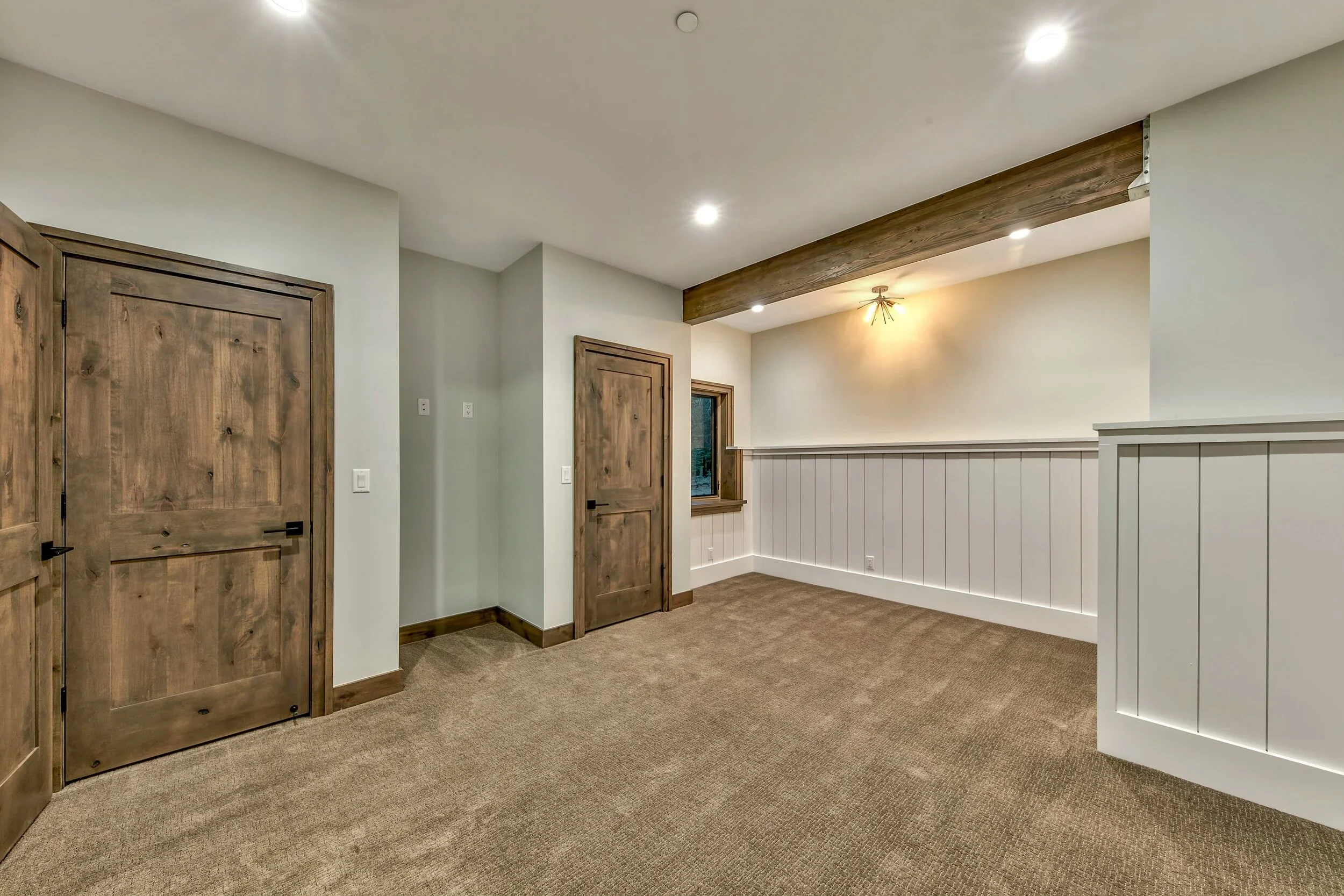 Empty room with beige carpet, white paneled half-walls, wooden doors, and a ceiling beam with recessed lighting.