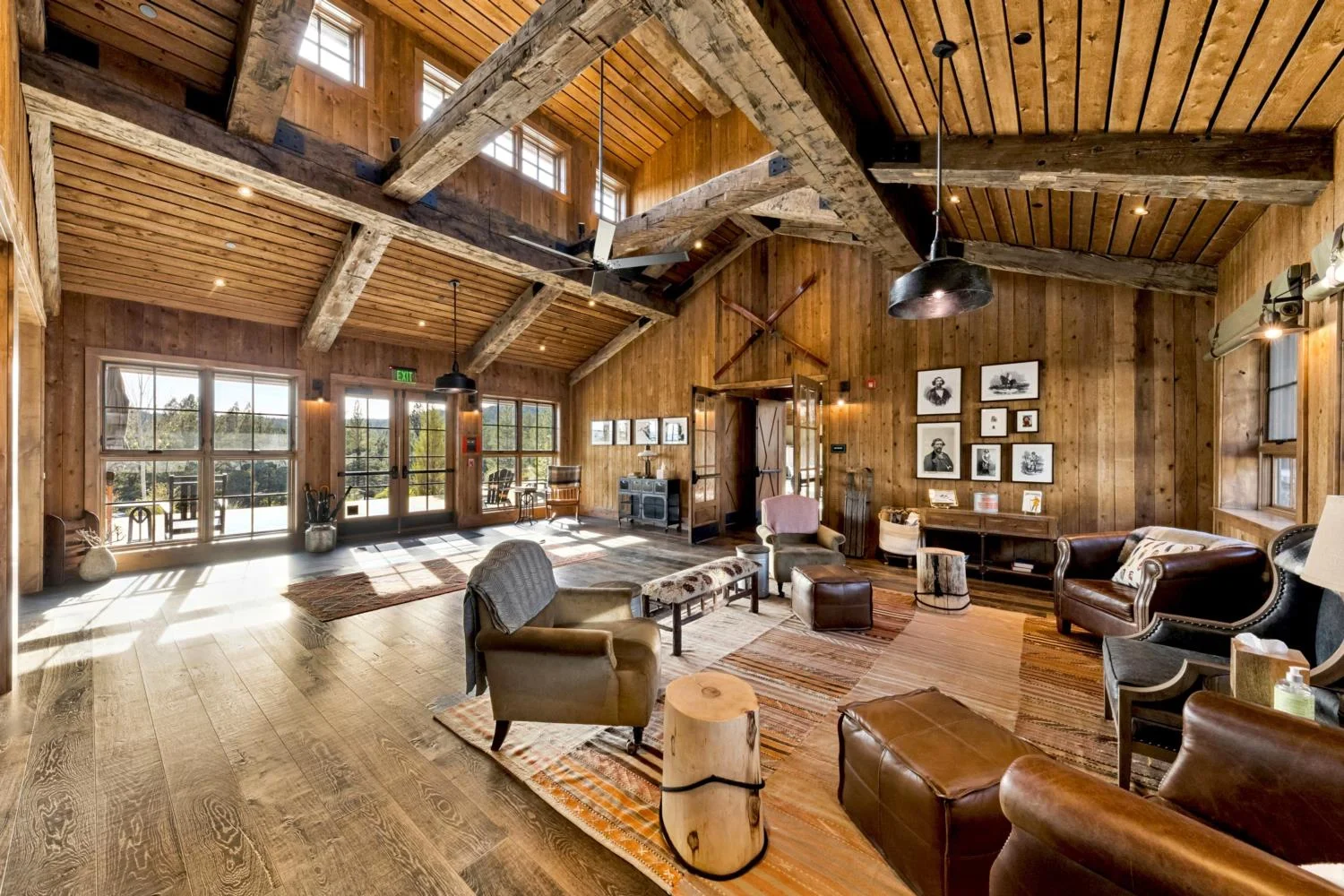 Spacious lodge lounge with dark wood flooring, reclaimed wood walls, a vaulted plank ceiling, exposed timber trusses, leather seating, patterned rugs, and large glass doors overlooking the landscape.