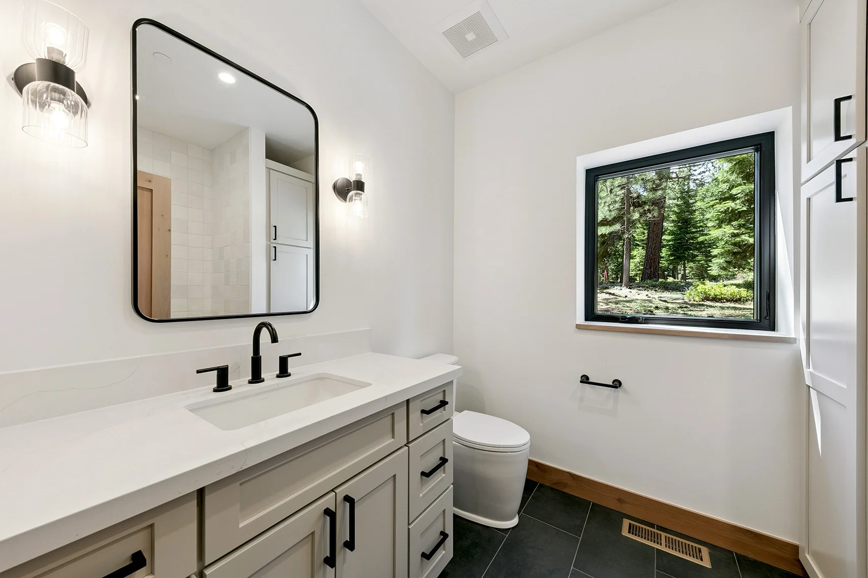 Modern white bathroom with a large mirror, black fixtures, and a window overlooking a peaceful pine tree setting.
