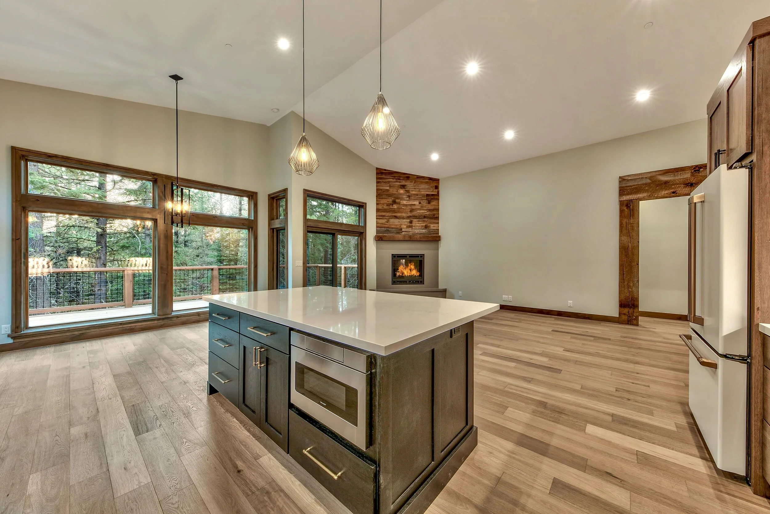 Open-concept kitchen and living room with large windows, island with white countertop, built-in microwave, fireplace, and wooden accents.