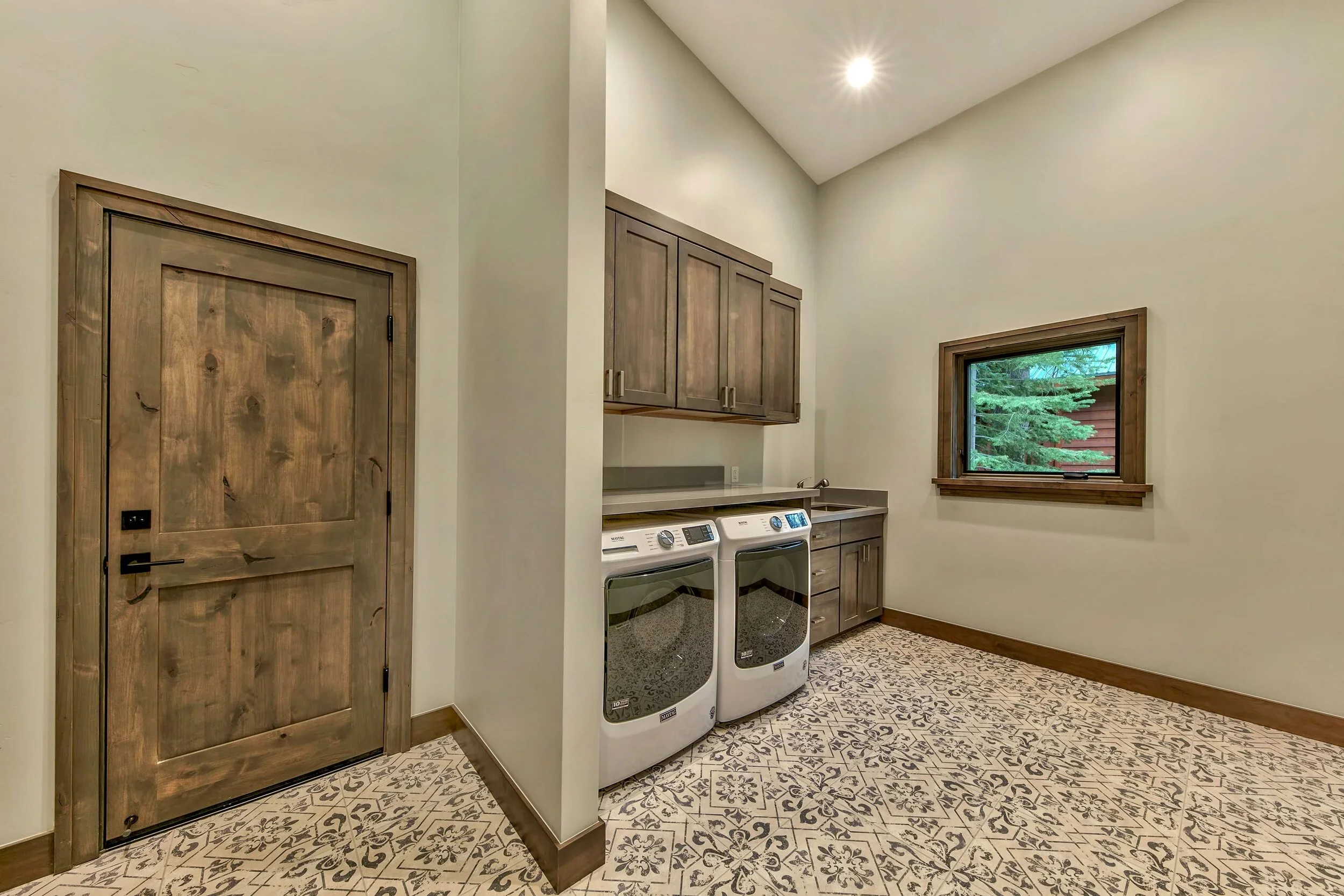 A laundry room with a wooden door, a window showing trees outside, a washing machine and dryer, wooden cabinets, and a patterned tile floor.