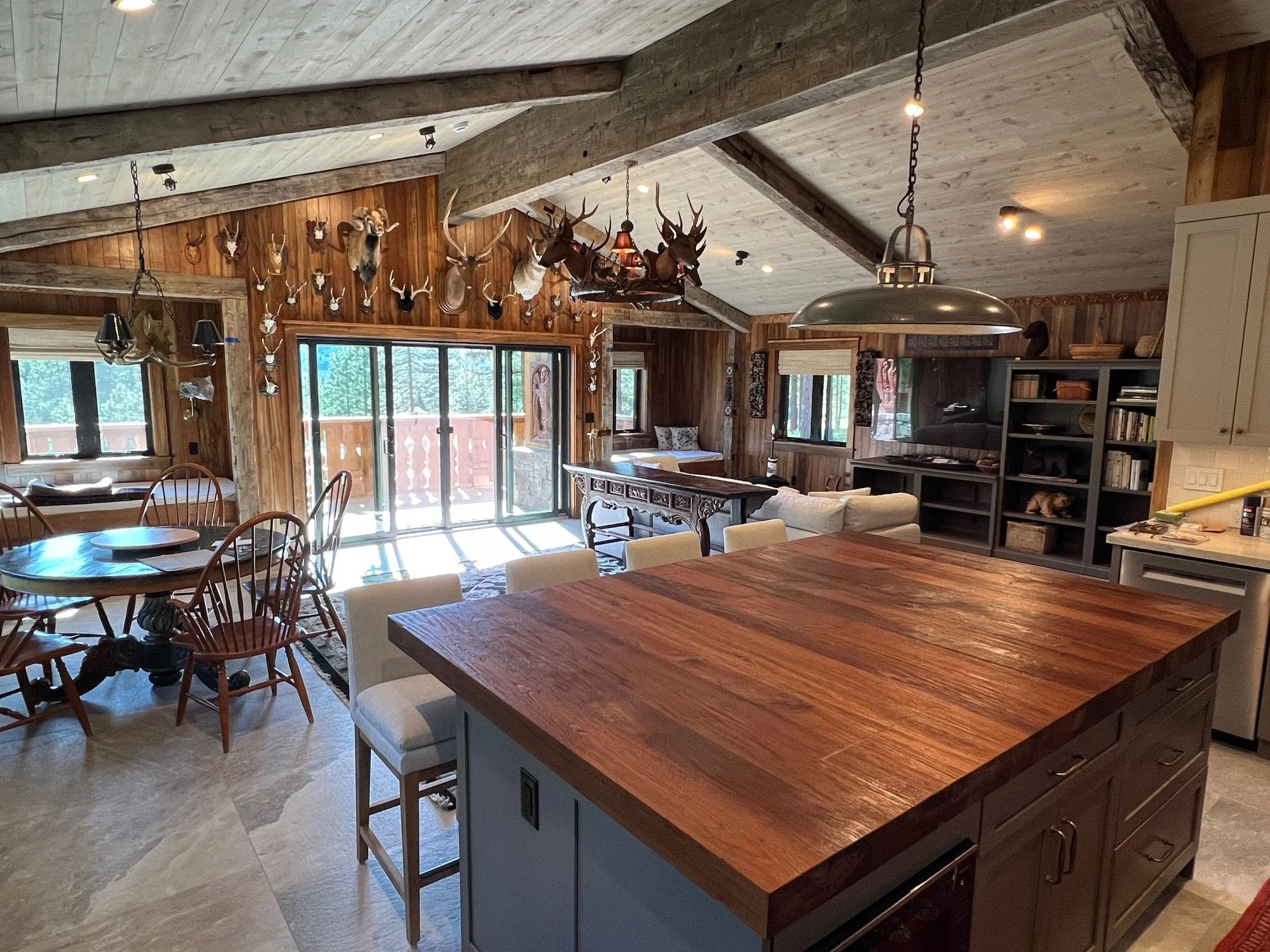 Interior of a rustic style home with a kitchen island, dining area, and living room. The decor includes mounted deer and antelope heads on the wooden walls and a large sliding glass door leading to a balcony. The ceiling is whitewashed wood with expo