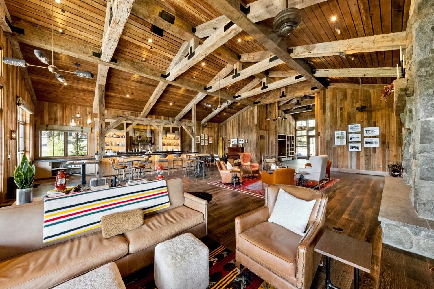Expansive lodge lounge with reclaimed wood walls, a vaulted plank ceiling, exposed timber trusses, leather seating, patterned rugs, a stone fireplace, and a full bar.