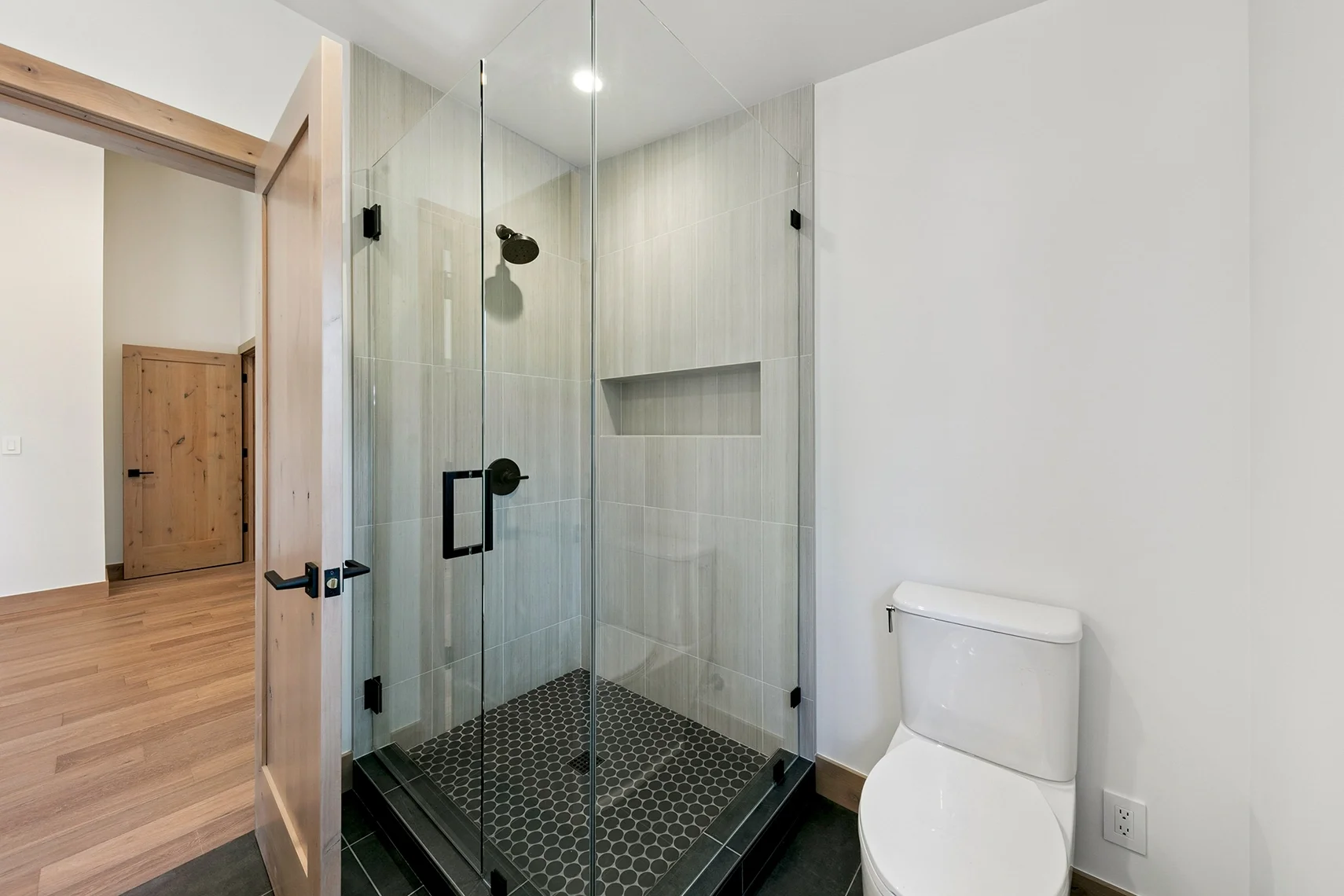Modern bathroom with white walls, white toilet, a glass-enclosed single shower, black penny tile shower flooring, and square black tile flooring throughout the space.