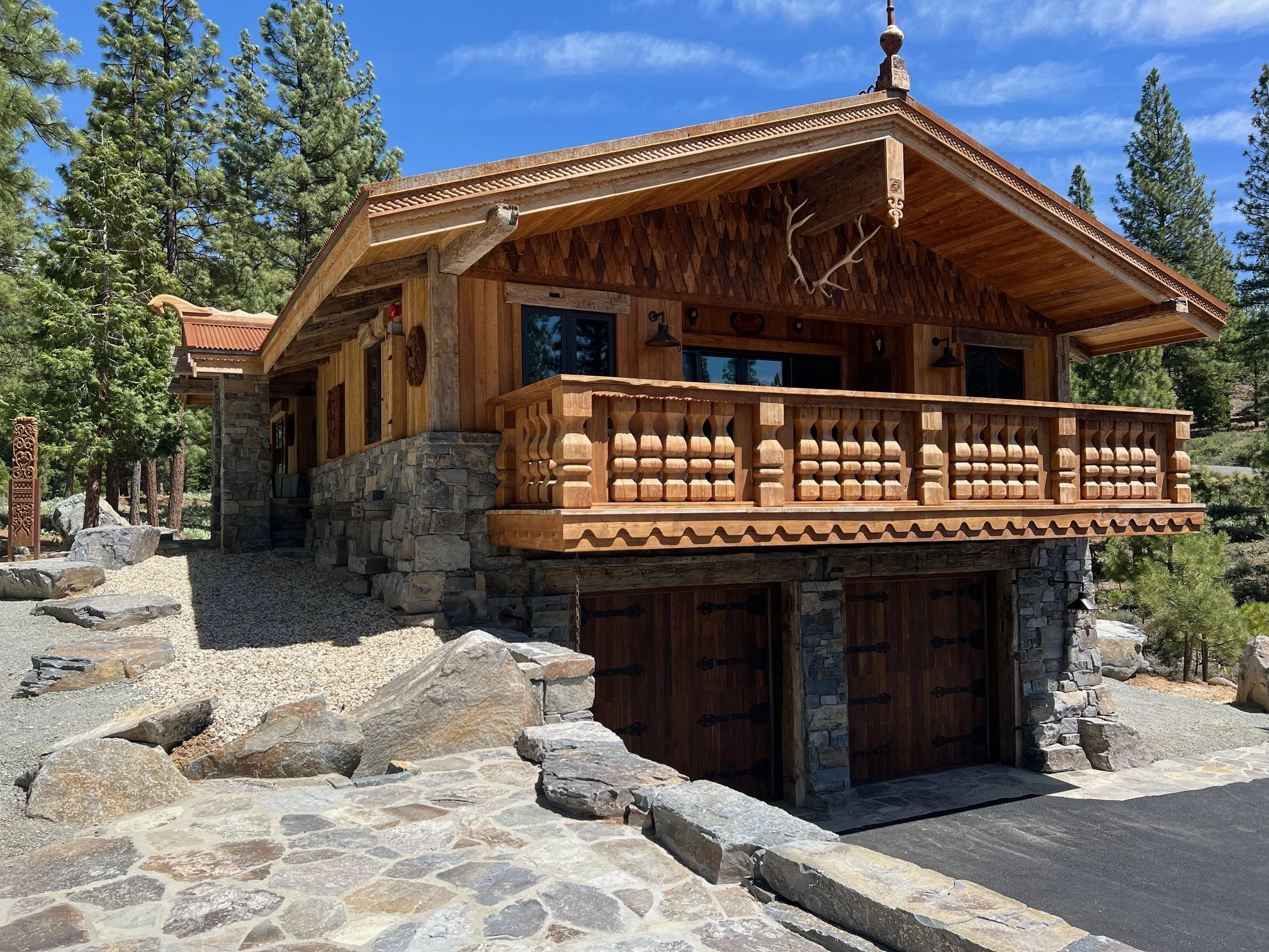 A rustic two-story mountain lodge with a wooden balcony, stone foundation, and a double garage with wooden doors, surrounded by trees and rocks.
