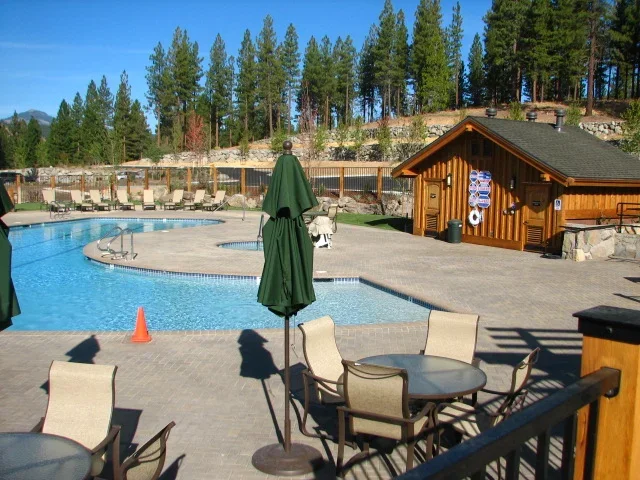 Poolside area with outdoor dining tables and chairs, a closed green umbrella, and a wooden pool house, with trees and a rocky hillside in the background on a sunny day.