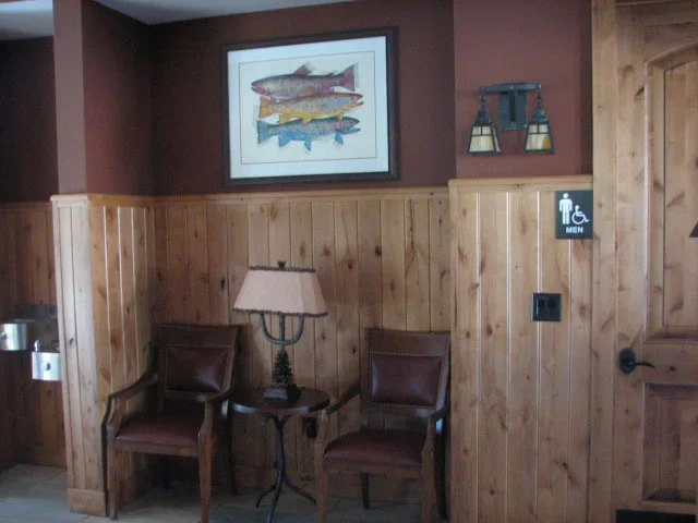 Waiting area with two wooden chairs with leather seats, a small side table with a lamp, a fish print on the wall, and a men's restroom sign.