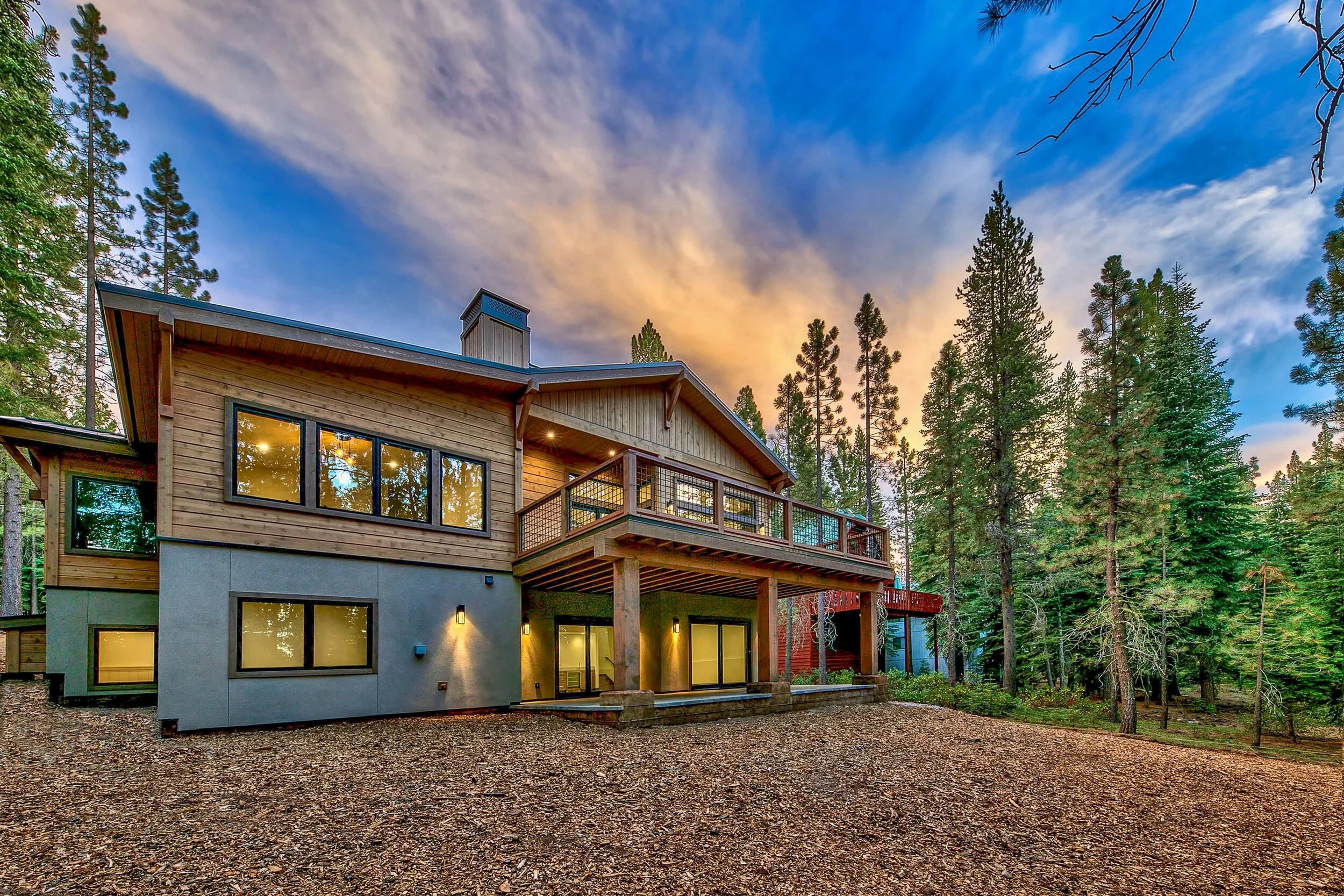 A modern two-story house with large windows, surrounded by tall pine trees, under a sunset sky with clouds.
