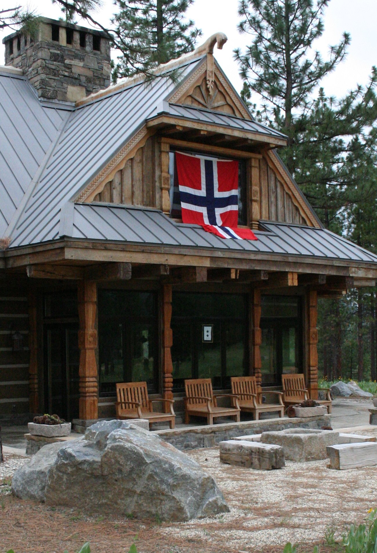 A rustic wooden house with a pine tree in the background, decorated with a Norwegian flag hanging from a second-story window.