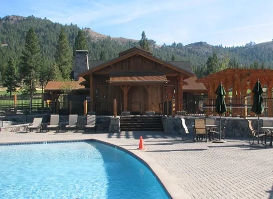 Pool area with lounge chairs, tables with green umbrellas, a wooden building, and a forested mountain in the background.
