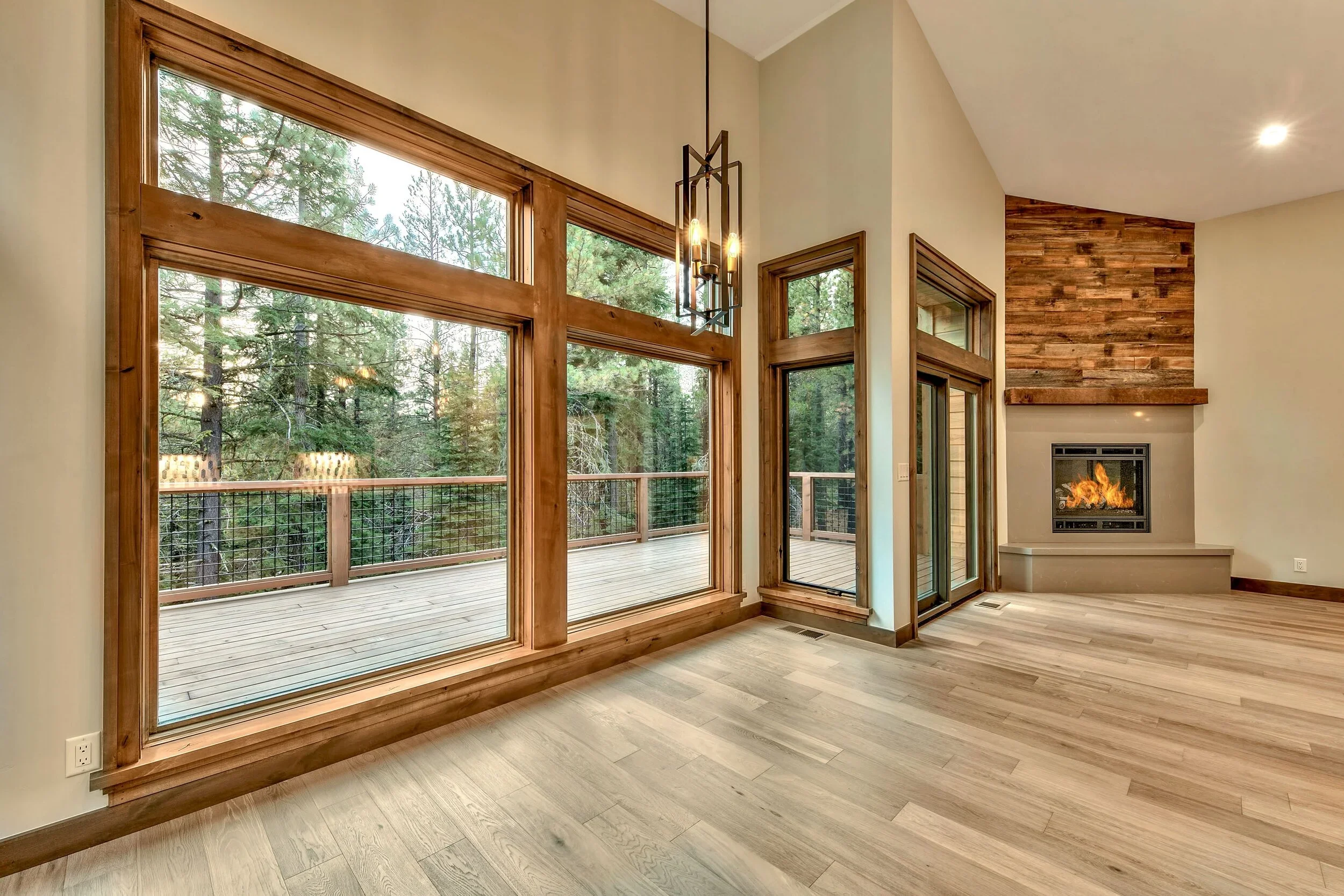Living room with large windows, a wooden deck outside, a ceiling light fixture, a fireplace with a wooden accent wall, and hardwood floors.