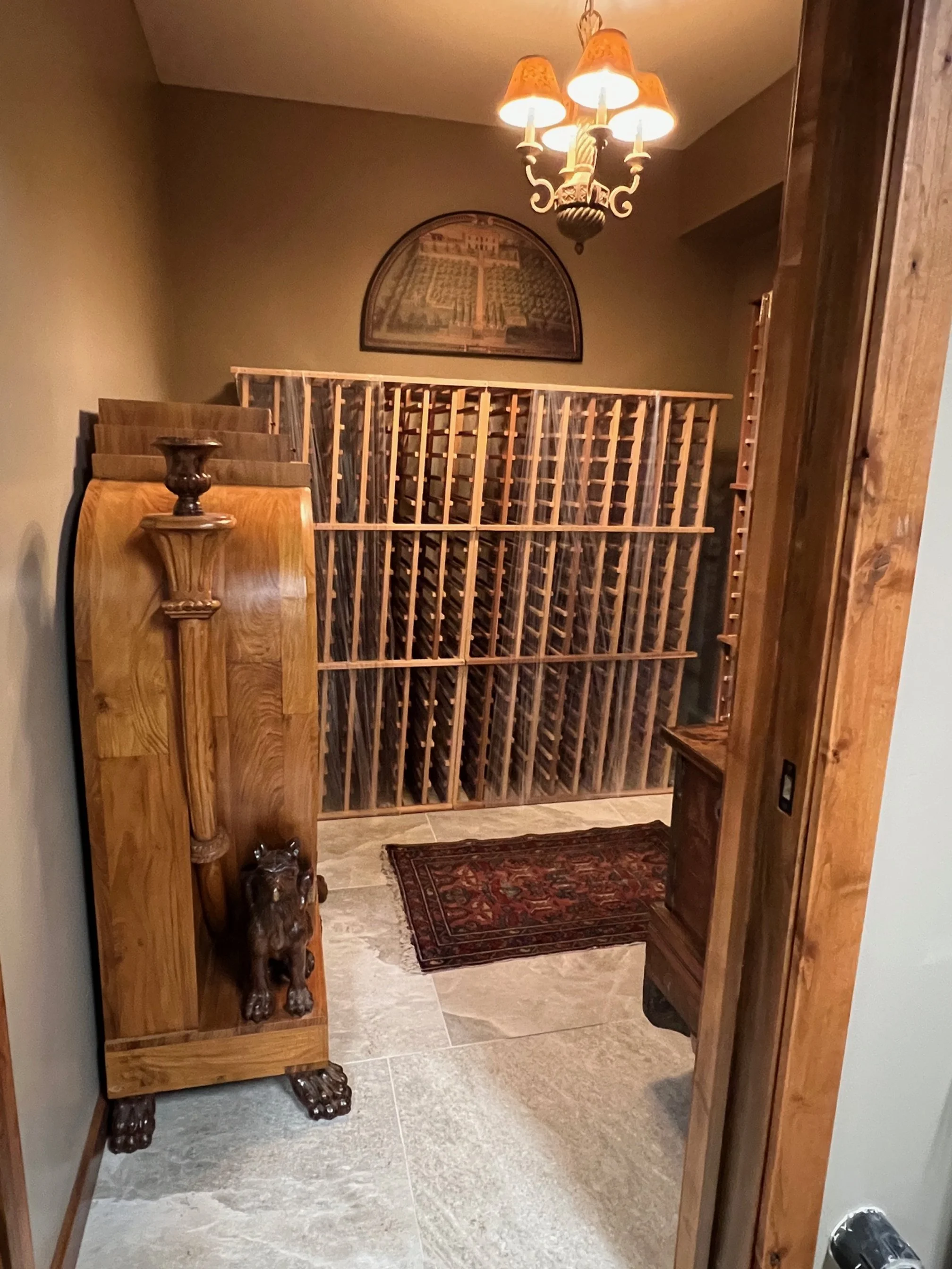 A wine cellar with wooden racks filled with wine bottles. Wooden furniture, a small rug, a wall art picture, and a chandelier with warm lighting.