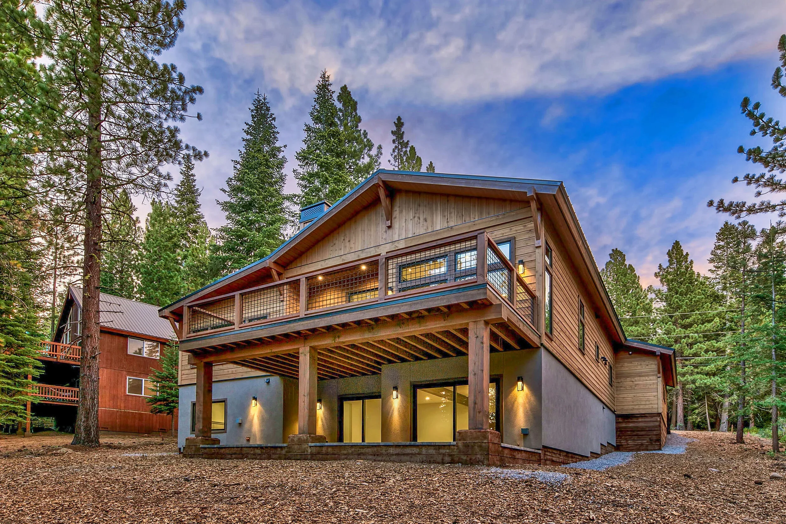 A modern house with a wooden exterior and large glass doors, situated among tall pine trees, with soft evening lighting.