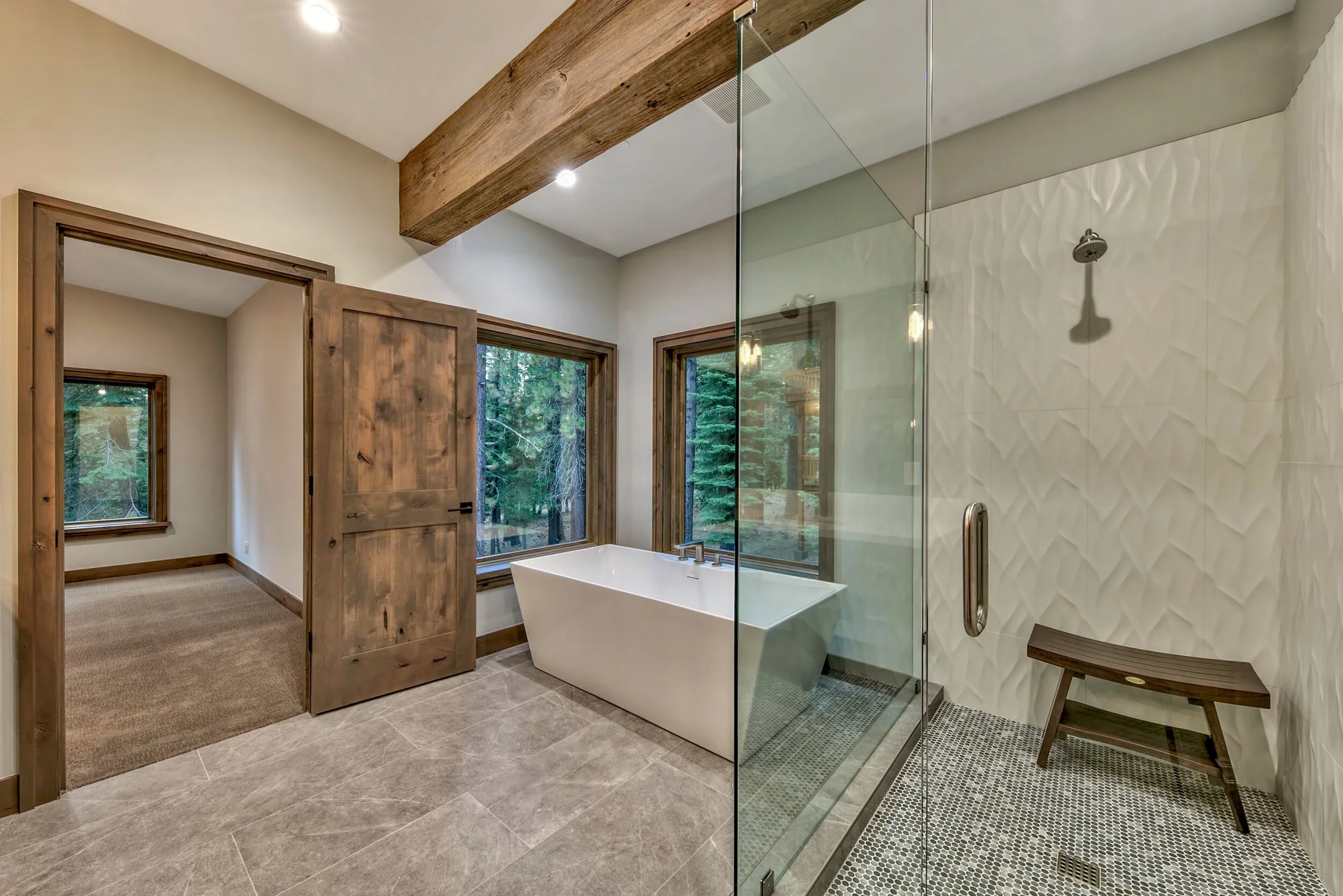 Modern bathroom with a freestanding bathtub, a glass-enclosed shower with a textured wall, wooden accents, and large windows showing greenery outside.