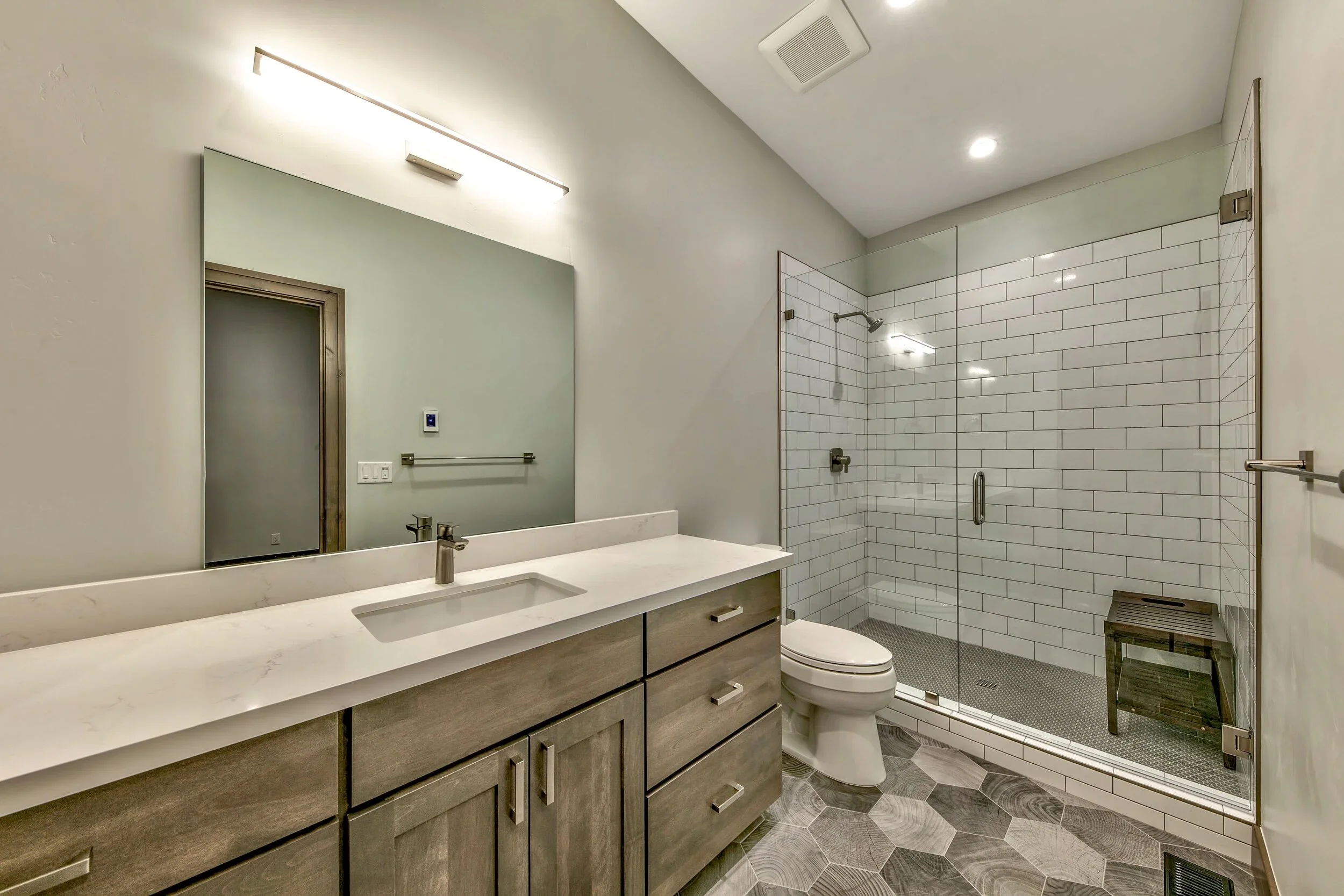 Modern bathroom with a large mirror, wooden vanity with a white countertop, a built-in sink, a wall-mounted faucet, a toilet, and a glass-enclosed shower with white subway tiles and a small wooden stool inside.