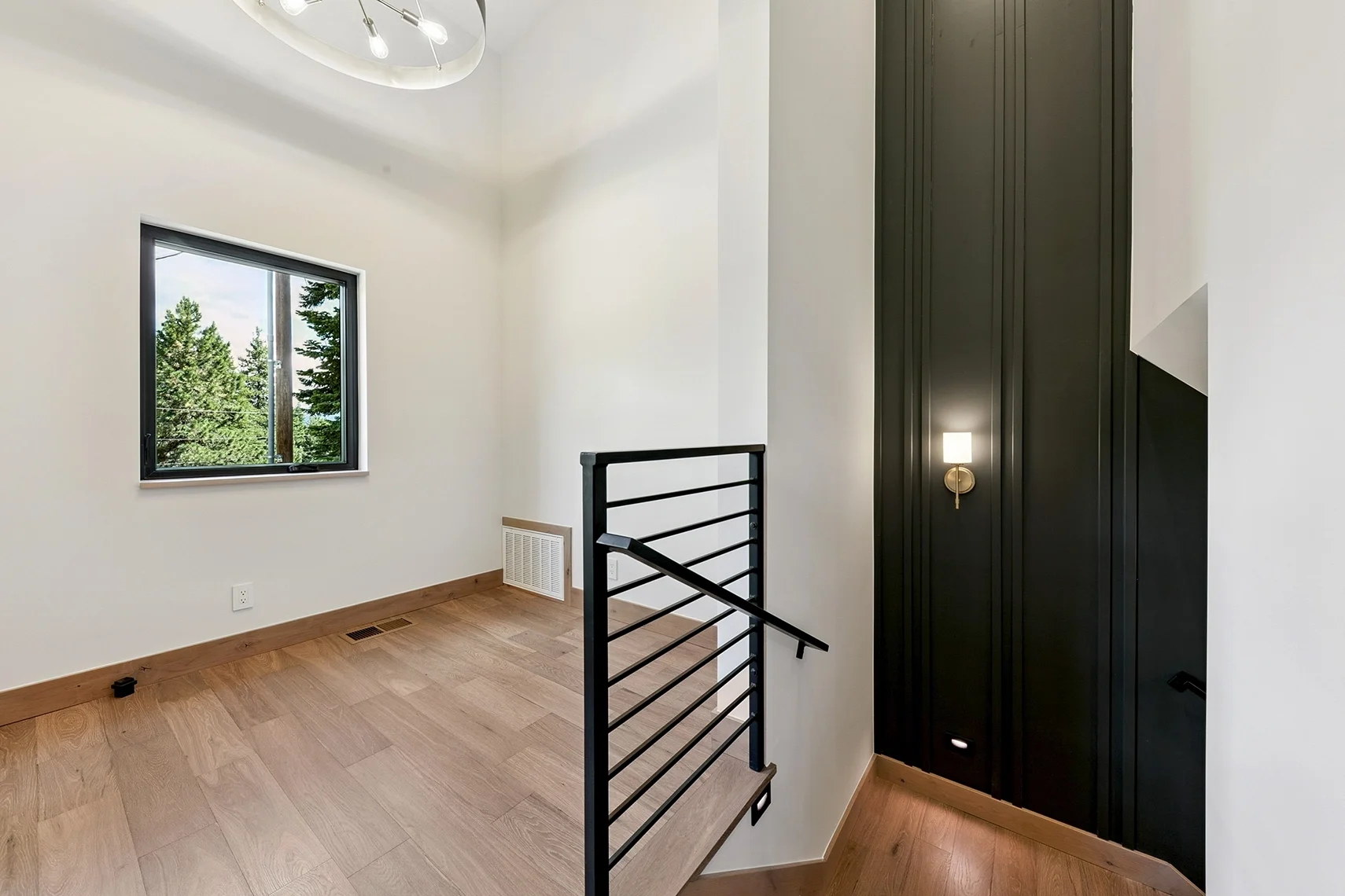 Modern entryway with wood flooring, a window, black wood-paneled accent wall, white walls, and an iron railing along the staircase leading downstairs.