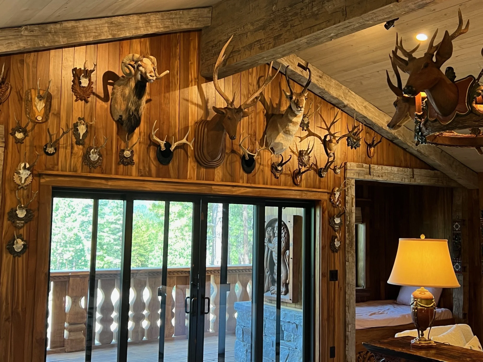 Interior of a rustic cabin with wood-paneled walls decorated with mounted animal heads, including deer and ram, near sliding glass doors leading to a balcony with a forest view, a cozy bedroom with a bed, and a table lamp.
