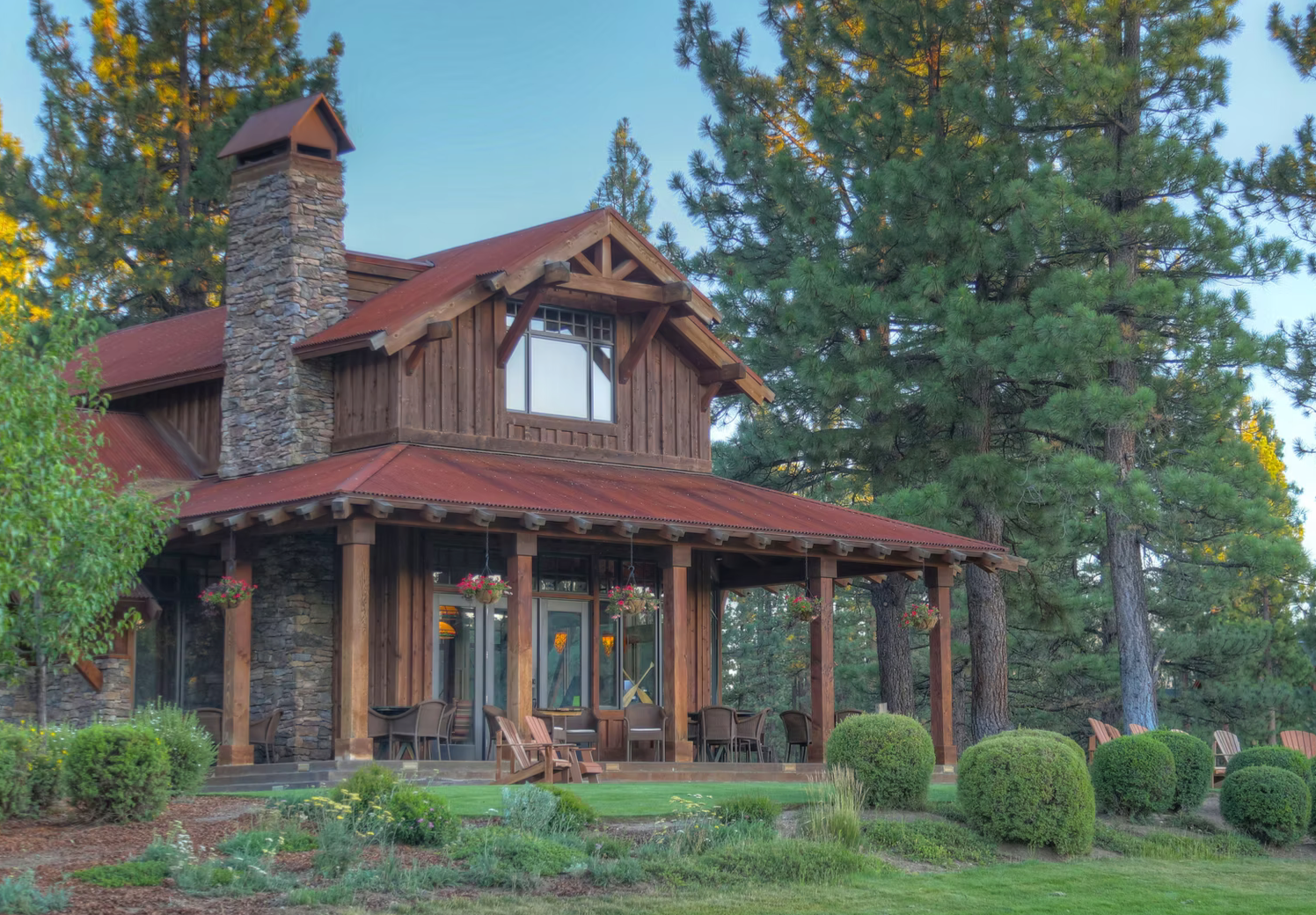 A rustic two-story house in a wooded area with a wraparound porch, large windows, hanging flower baskets, and a well-maintained lawn with bushes and trees.