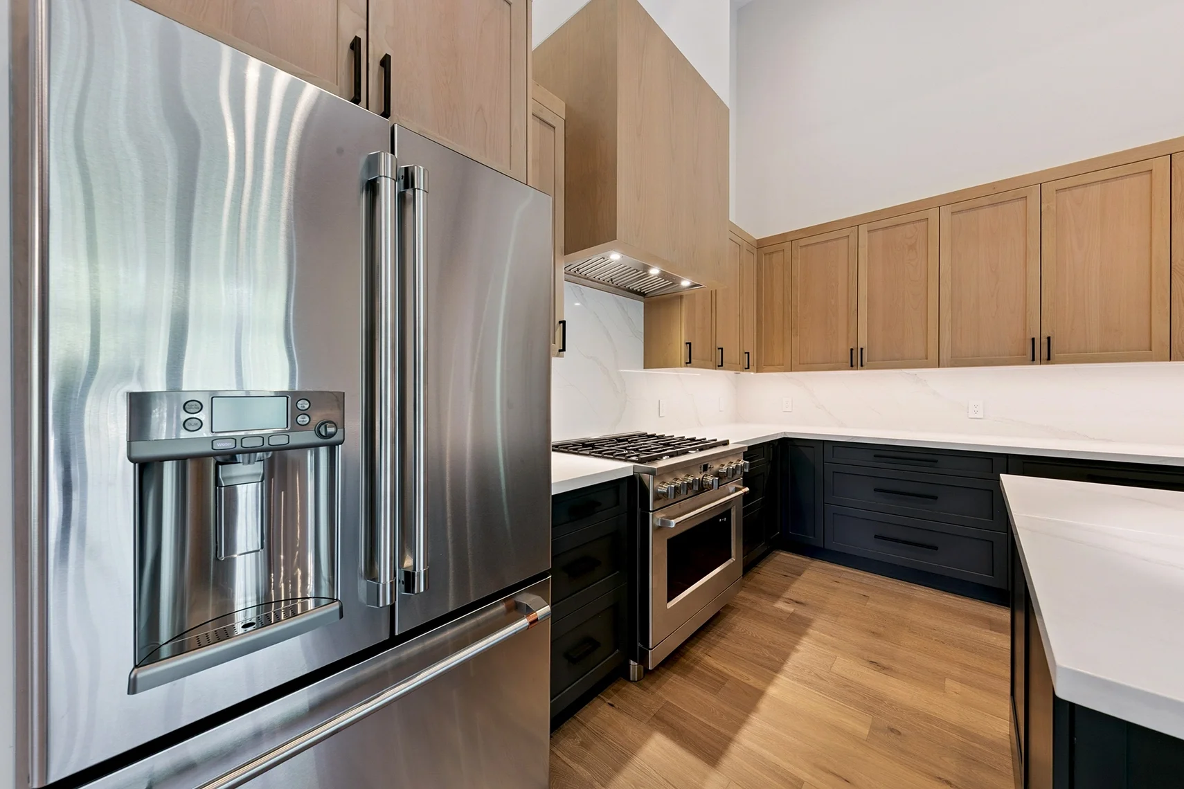 Modern kitchen with a large stainless steel refrigerator, natural wood upper cabinets, black lower cabinets, and white backsplash and countertops.
