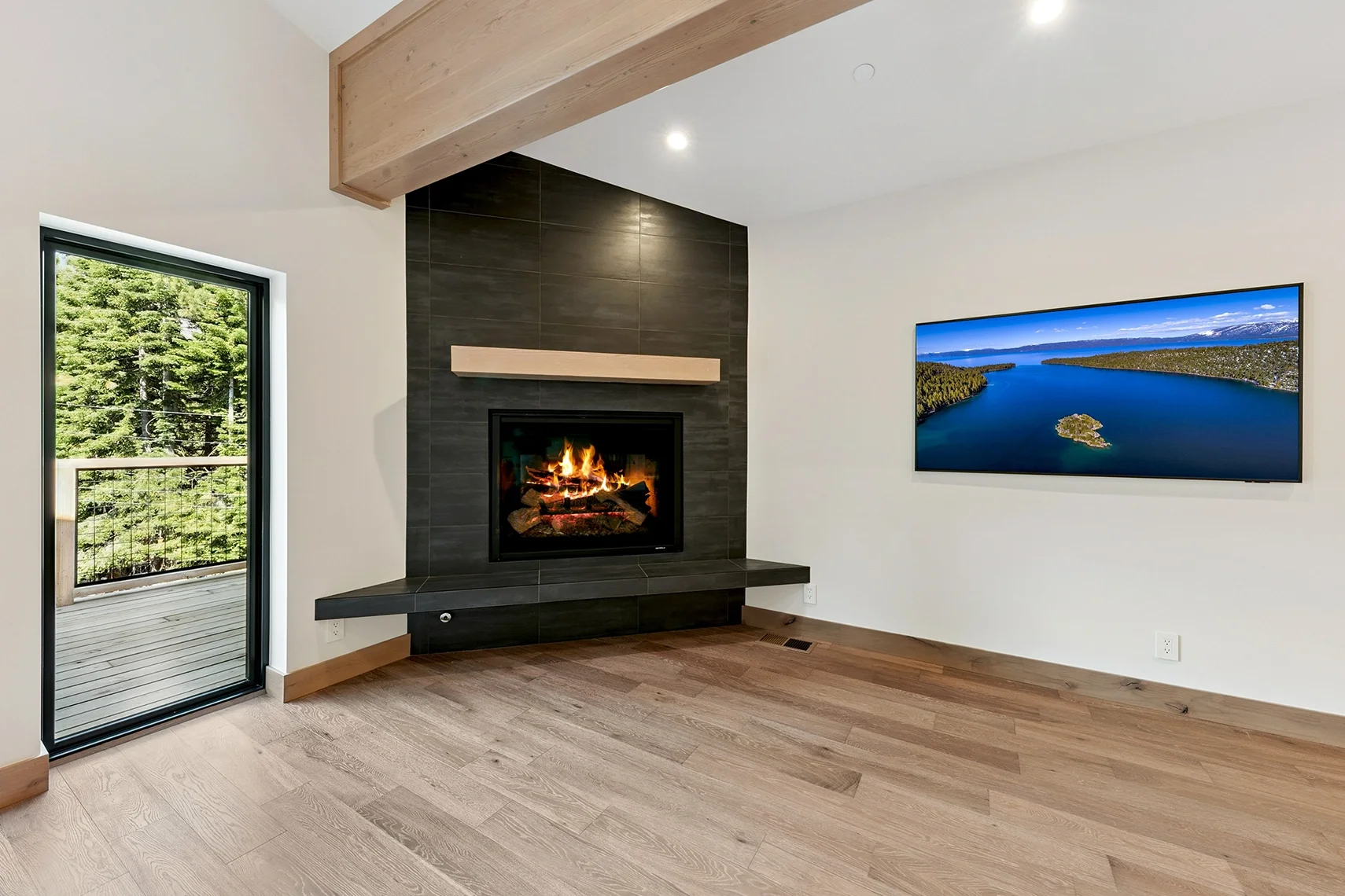 Modern living room with a large black fireplace, natural wood flooring, white walls, exposed ceiling beams, a wall-mounted TV, and a door opening to the deck.